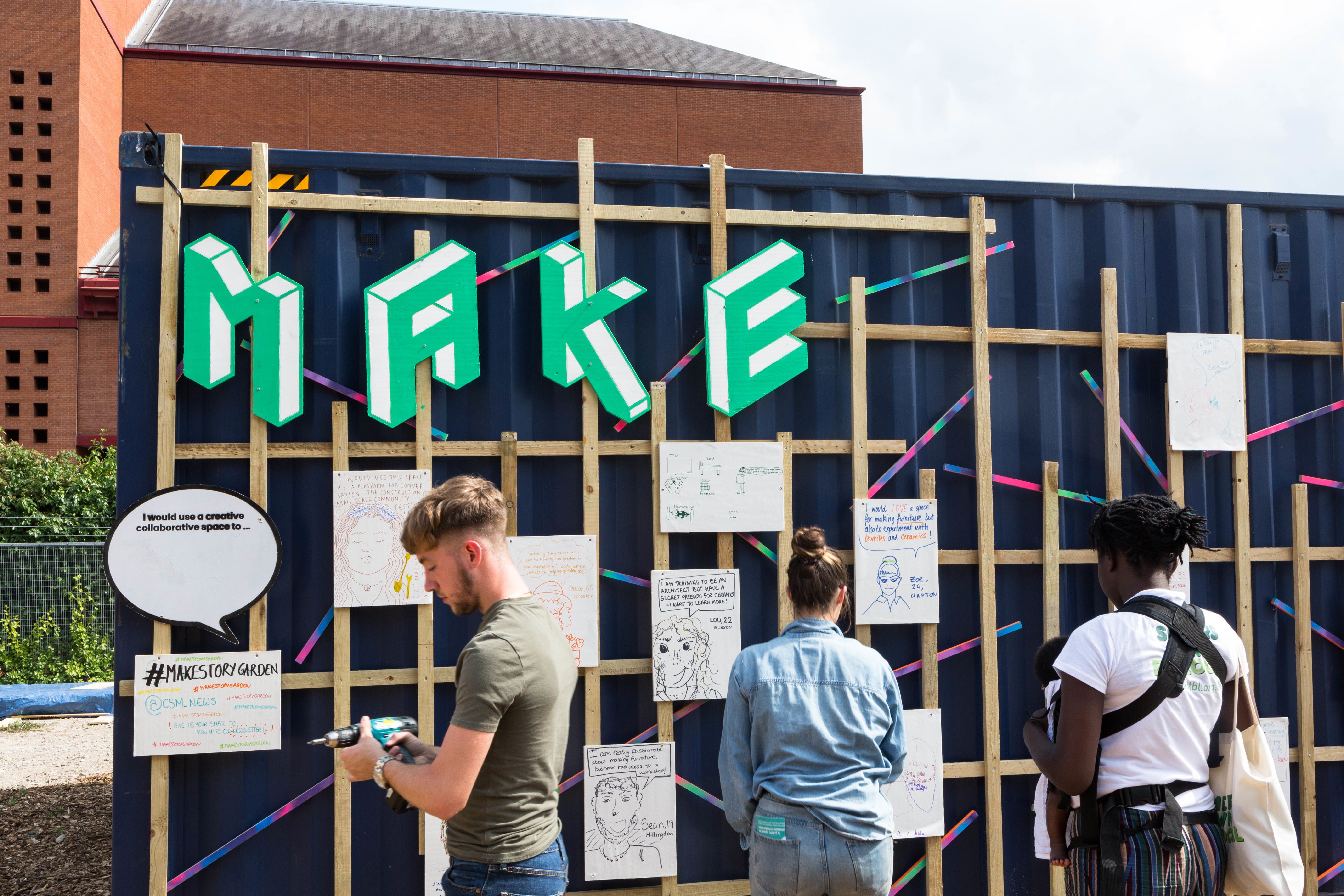 People in front of a shipping container with the letters MAKE in green caps and there is a trellis with hanging sheets of A4 and they are writing on some of them