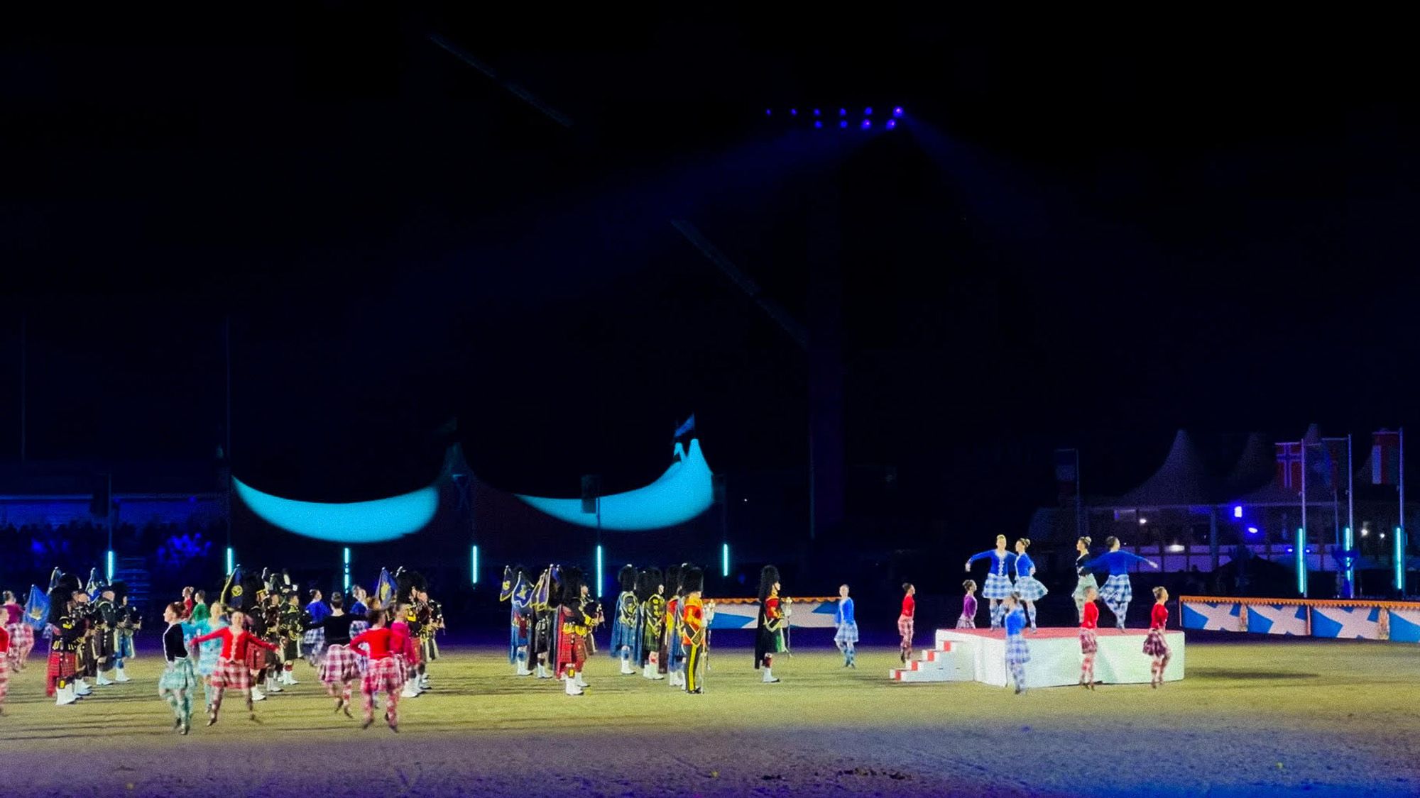 It is dark. At the centre of a large arena, Highland dancers perform a ceilidh. Dressed in blue and red, they dance around a formation of pipers playing the bagpipes. Partially illuminated tents and the crowd can be seen in the background.
