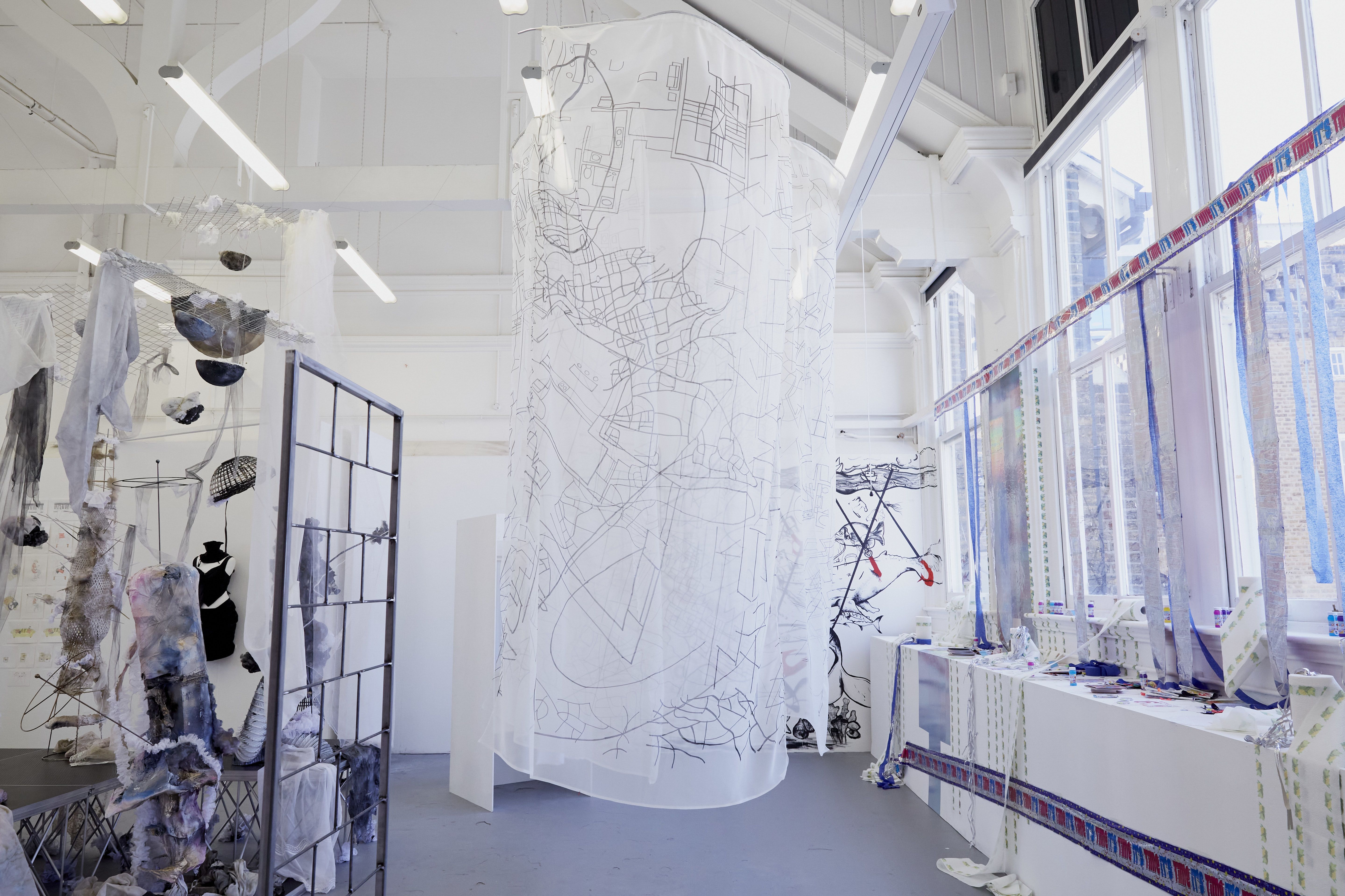 Image shows a white sheet of fabric hanging from the ceiling with a metal frame and different coloured fabric to the left and a wall of windows to the right. The white sheet of fabric has drawings all over it. The room that it is in is painted white with a blue-grey floor.