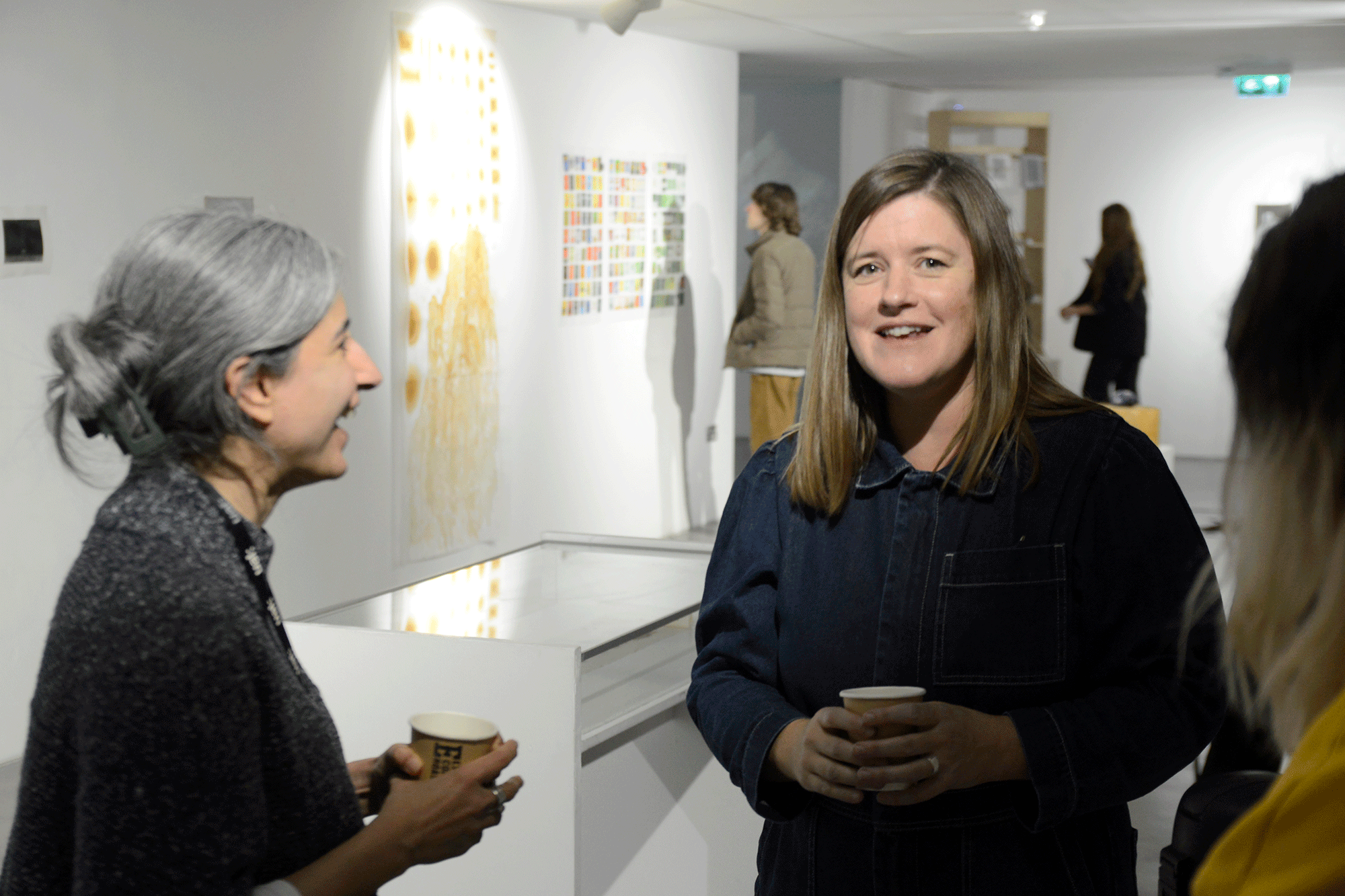 A person looks towards the camera holding a cup, while another person looks towards them. In the background, you can see other people looking at artwork on the walls.