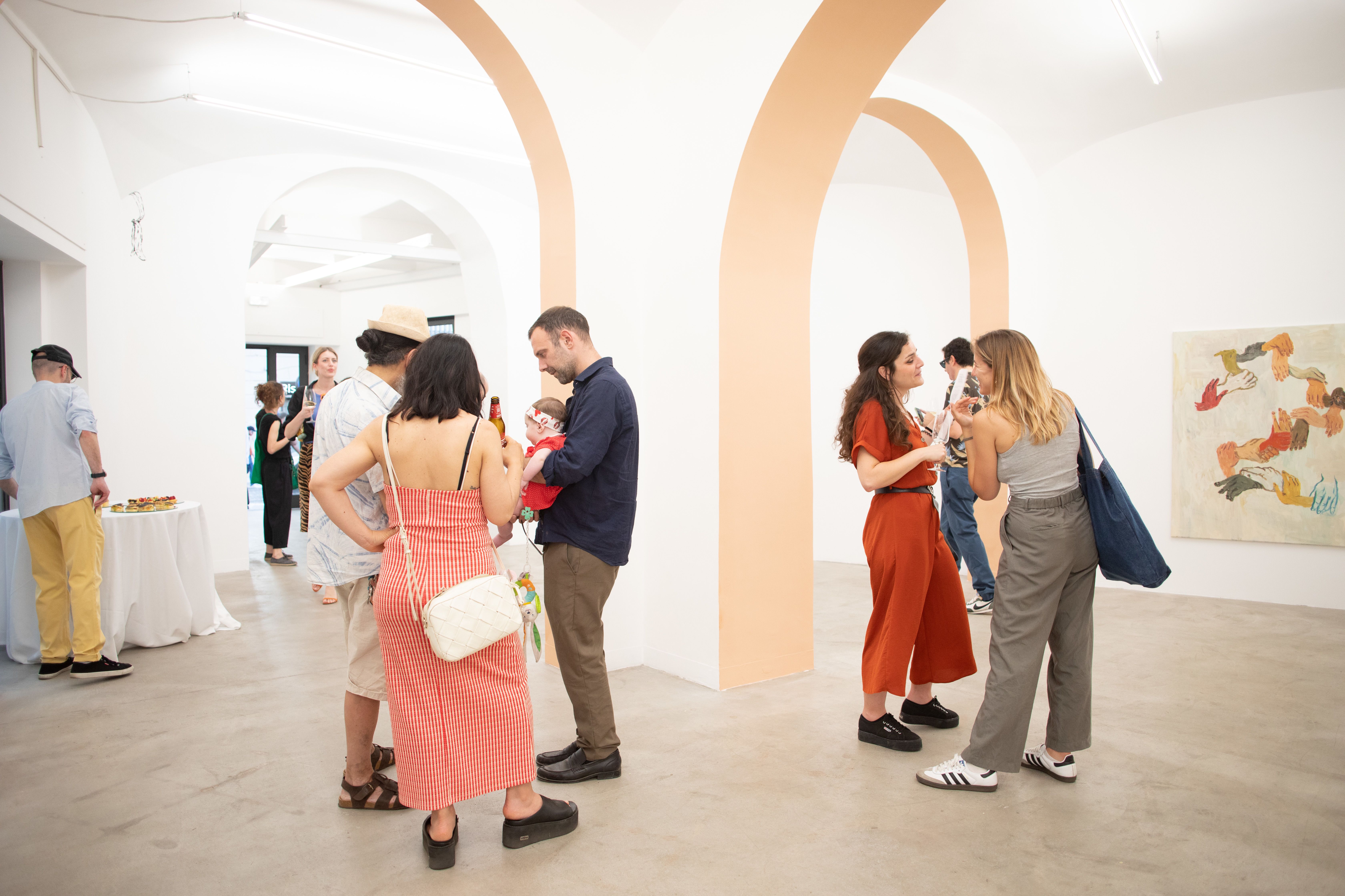 People standing in the gallery, a series of rooms connected by large arches. The room is bright, painted white with peach colour on the underside of the arches. 