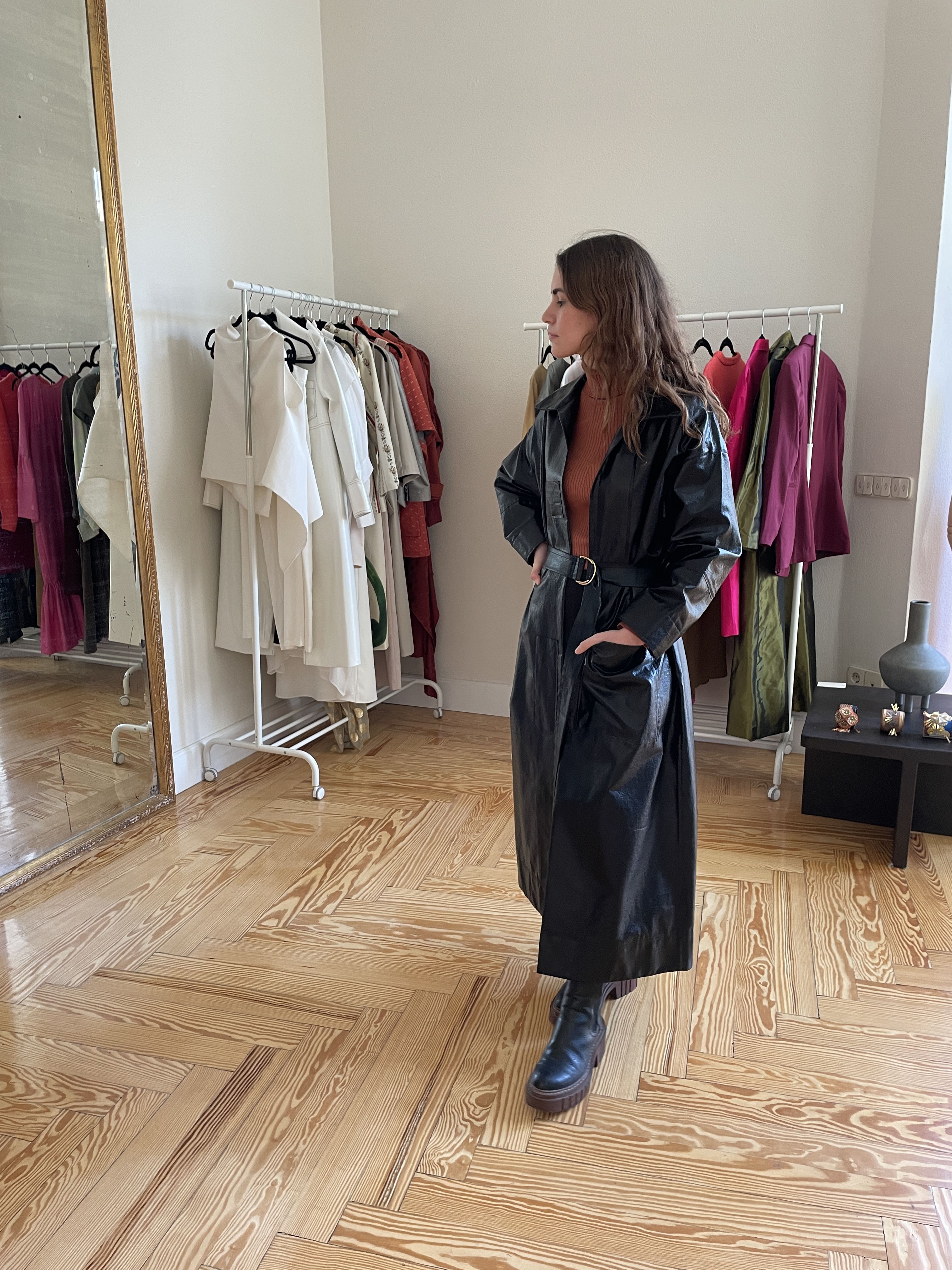 Woman in long black leather-look trenchcoat, standing in front of a full length mirror with 2 clothes rails behind her. 