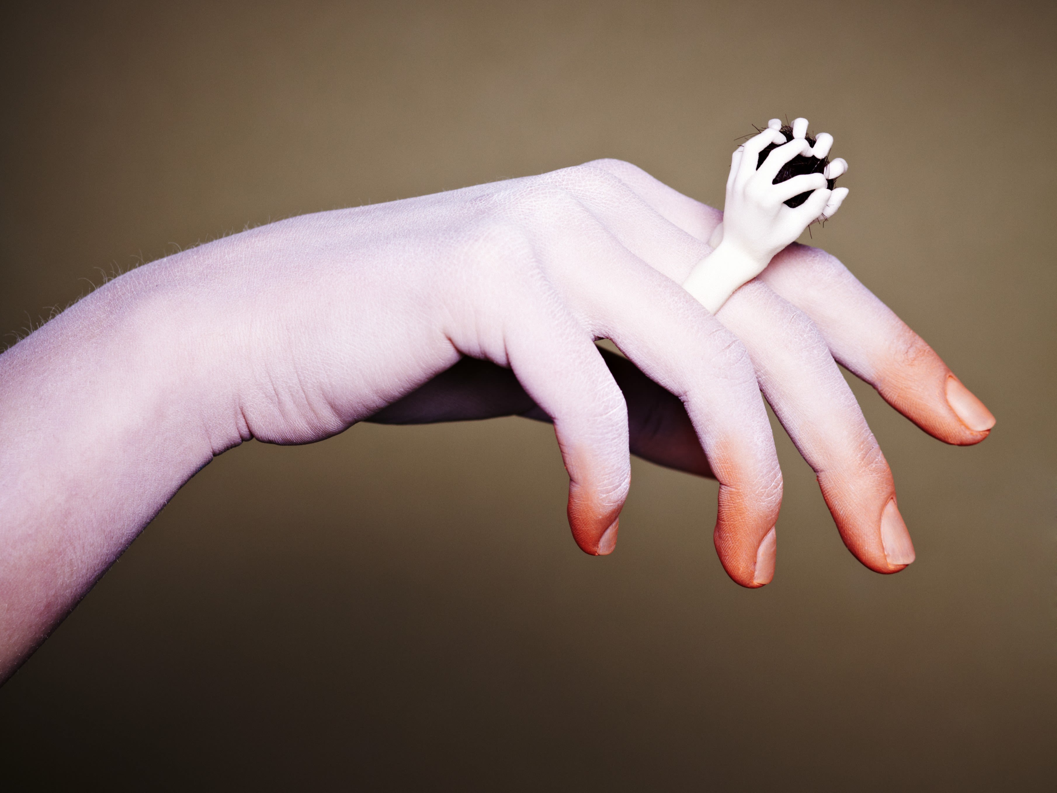 A large pale hand, stretched out against a muted brown background, has a tiny white arm gripping its index finger. The image feels surreal and intriguing.