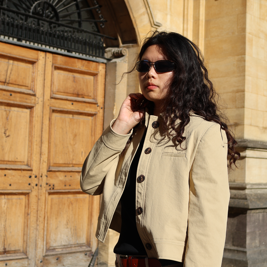 A student wearing sunglasses and a light jacket stands outdoors in front of a stone building and wooden doors, adjusting their hair in sunlight.