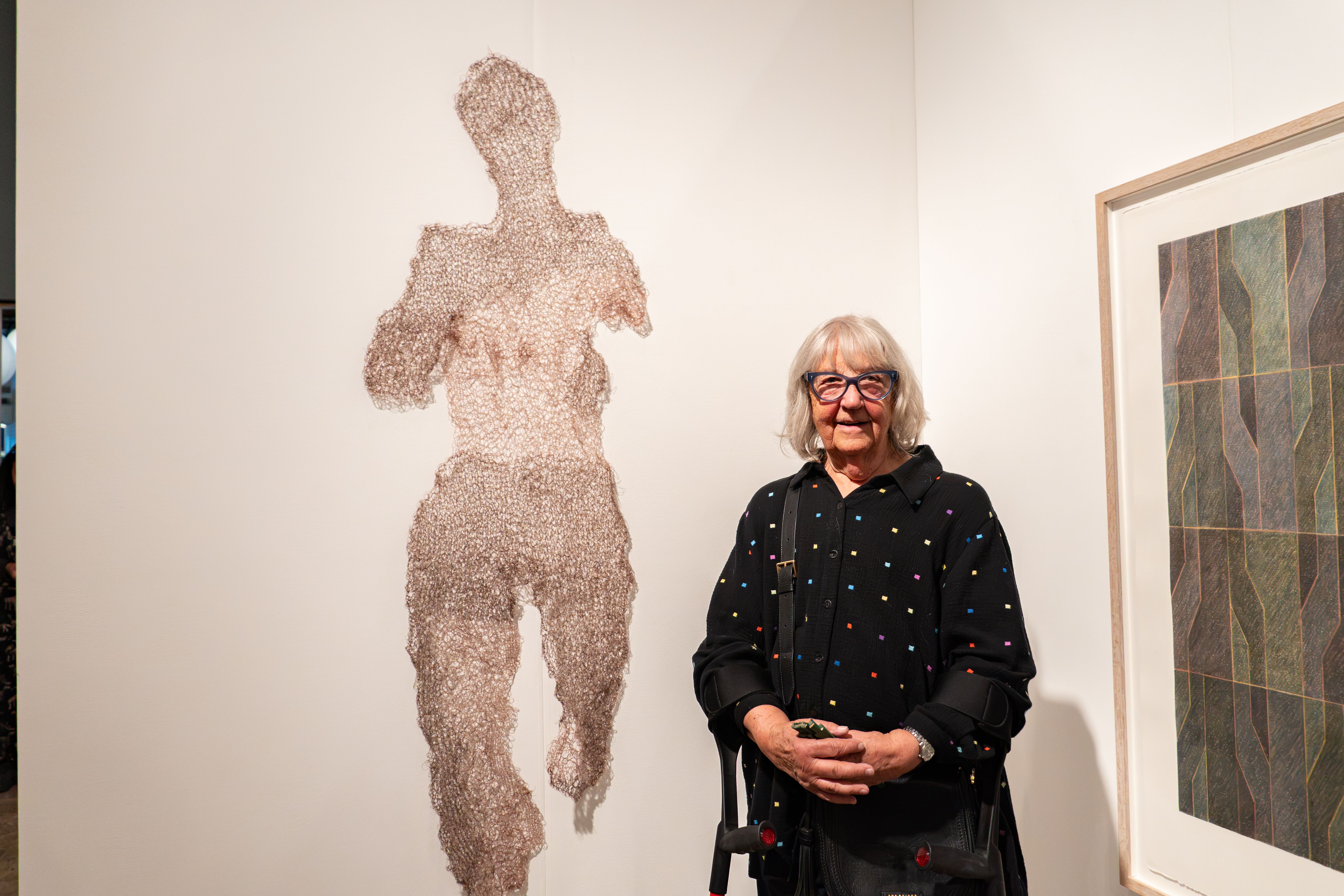 Yvonne stands in an art gallery beside a large, human-shaped wall sculpture made from delicate wire or mesh. Framed abstract artwork hangs on the wall to the right. The gallery is brightly lit with neutral walls, highlighting the texture of the sculpture and the geometric patterns in the framed piece.
