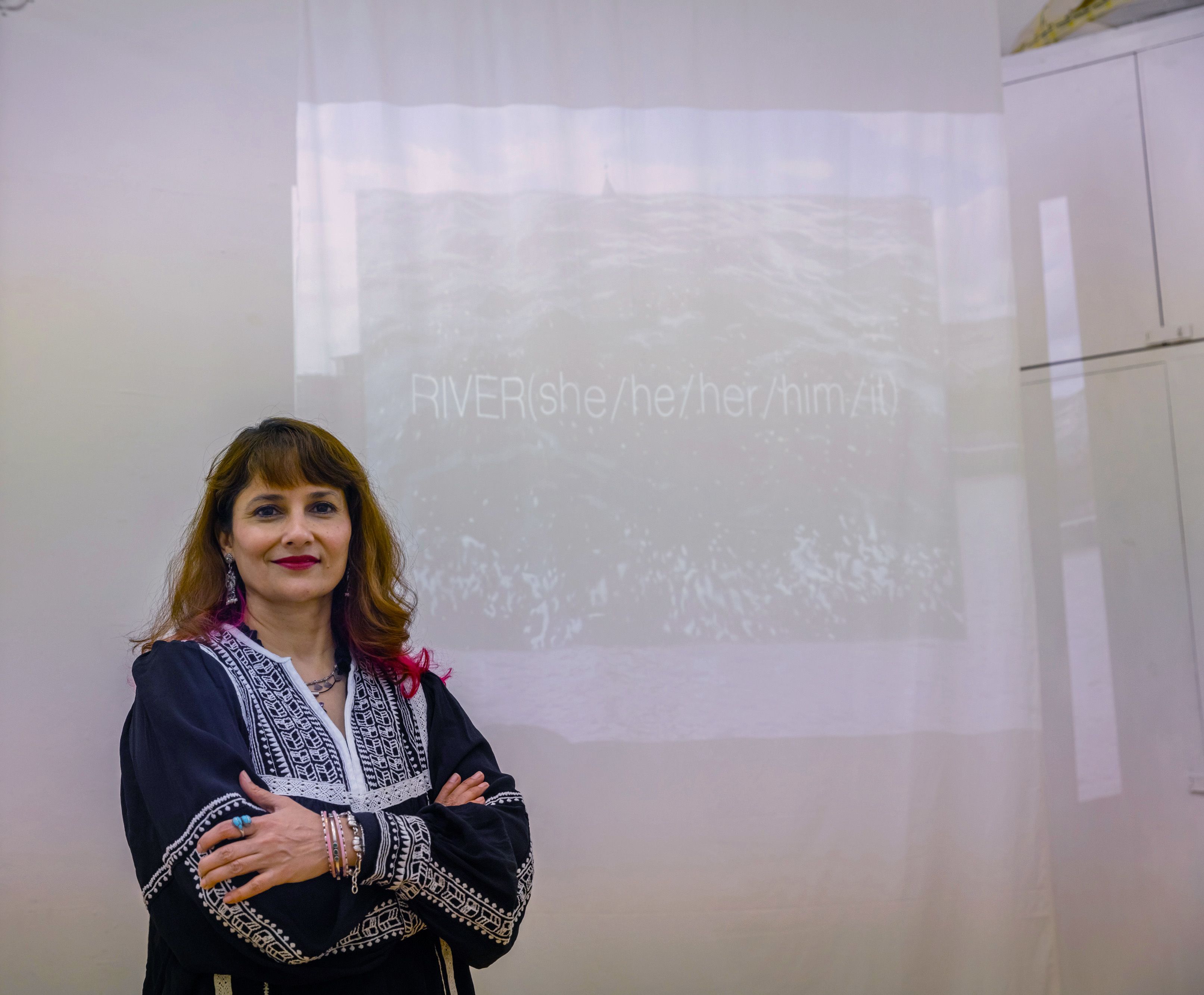 Anita stood infront of a projection that reads 'RIVER (she/he/him/it)' 