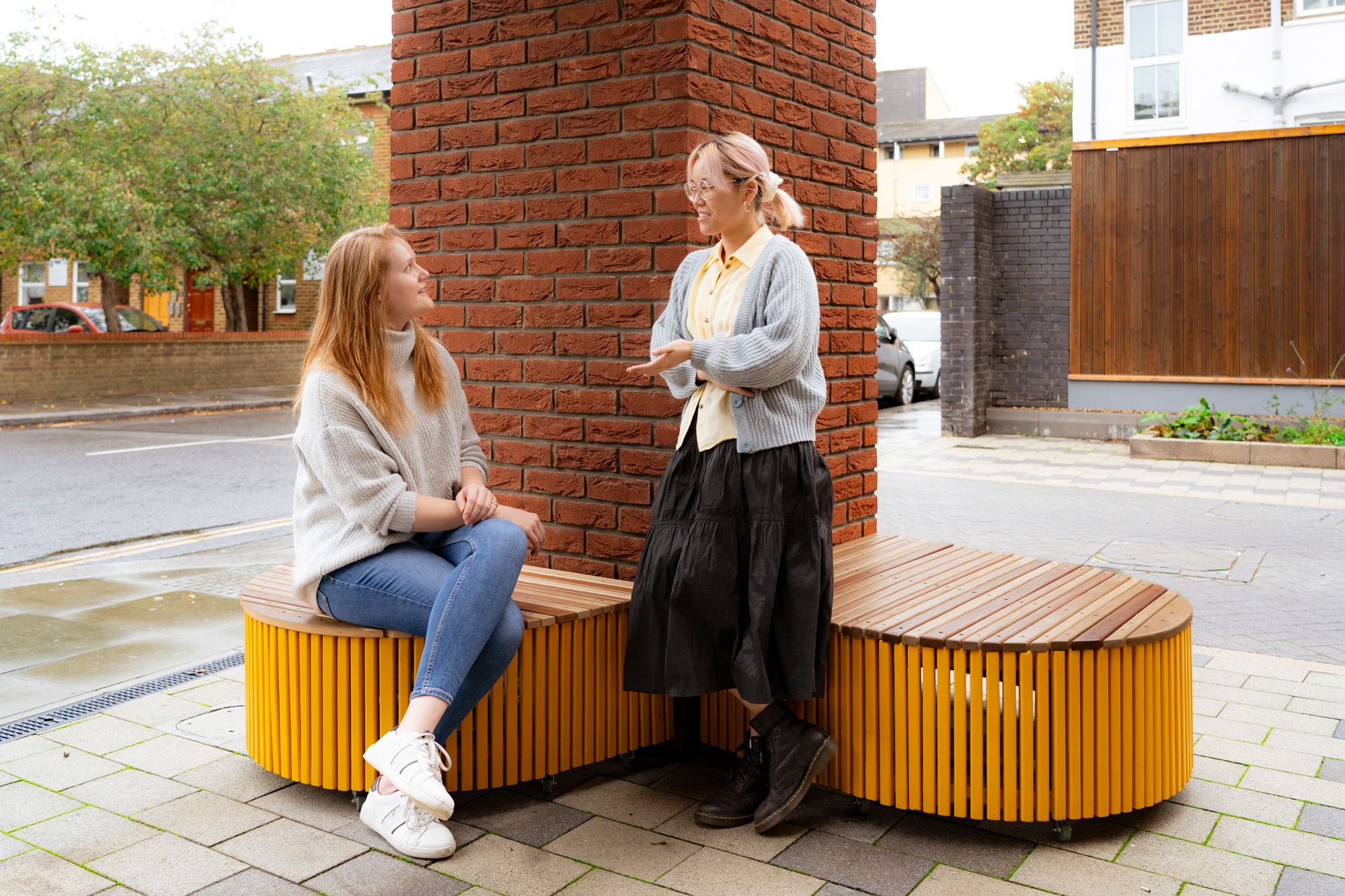 Two woman are talking, one is seated on a bench while the other is leaning against a brick wall
