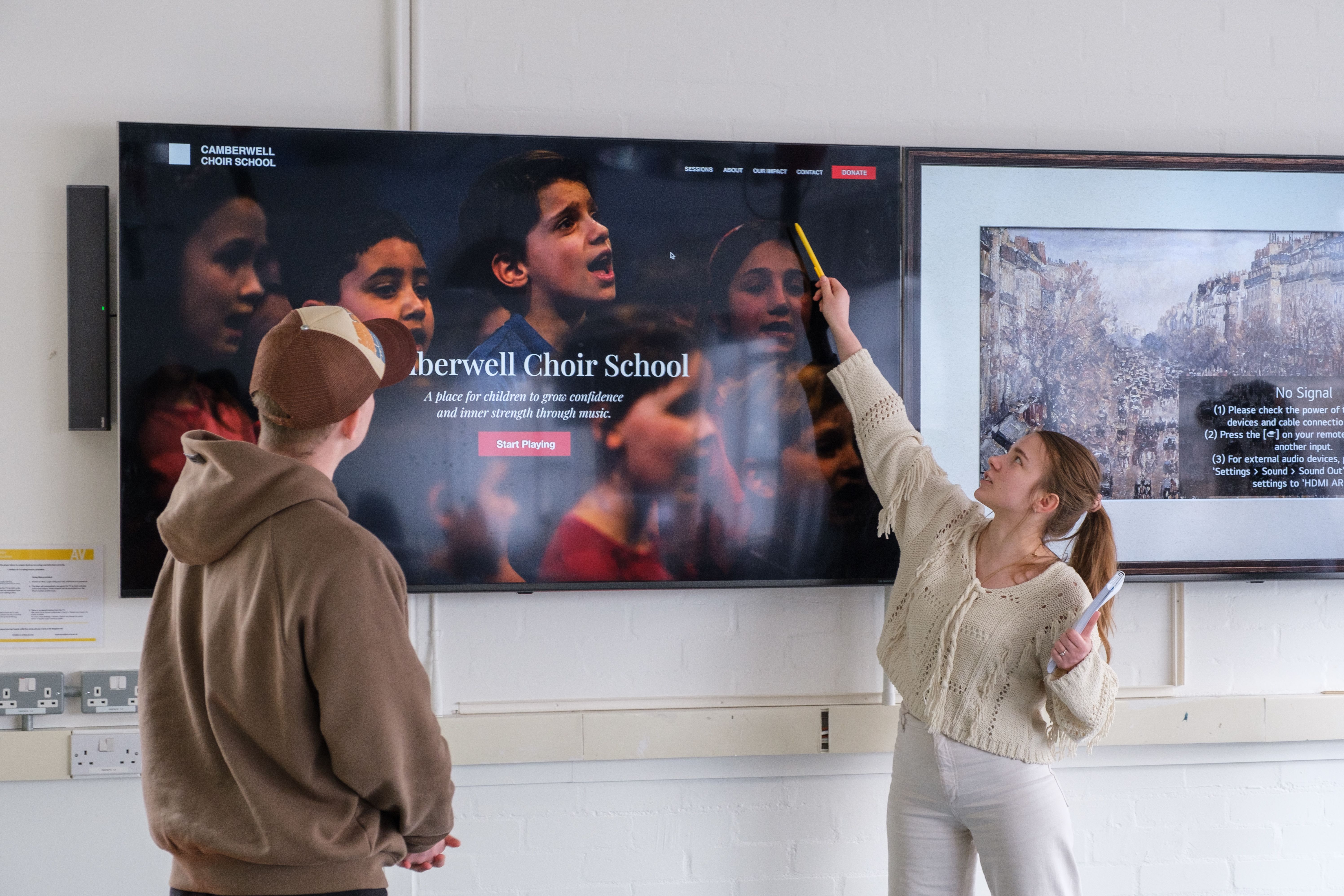 Two students present their website design, one points at a screen on the wall and the other looks in that direction too.