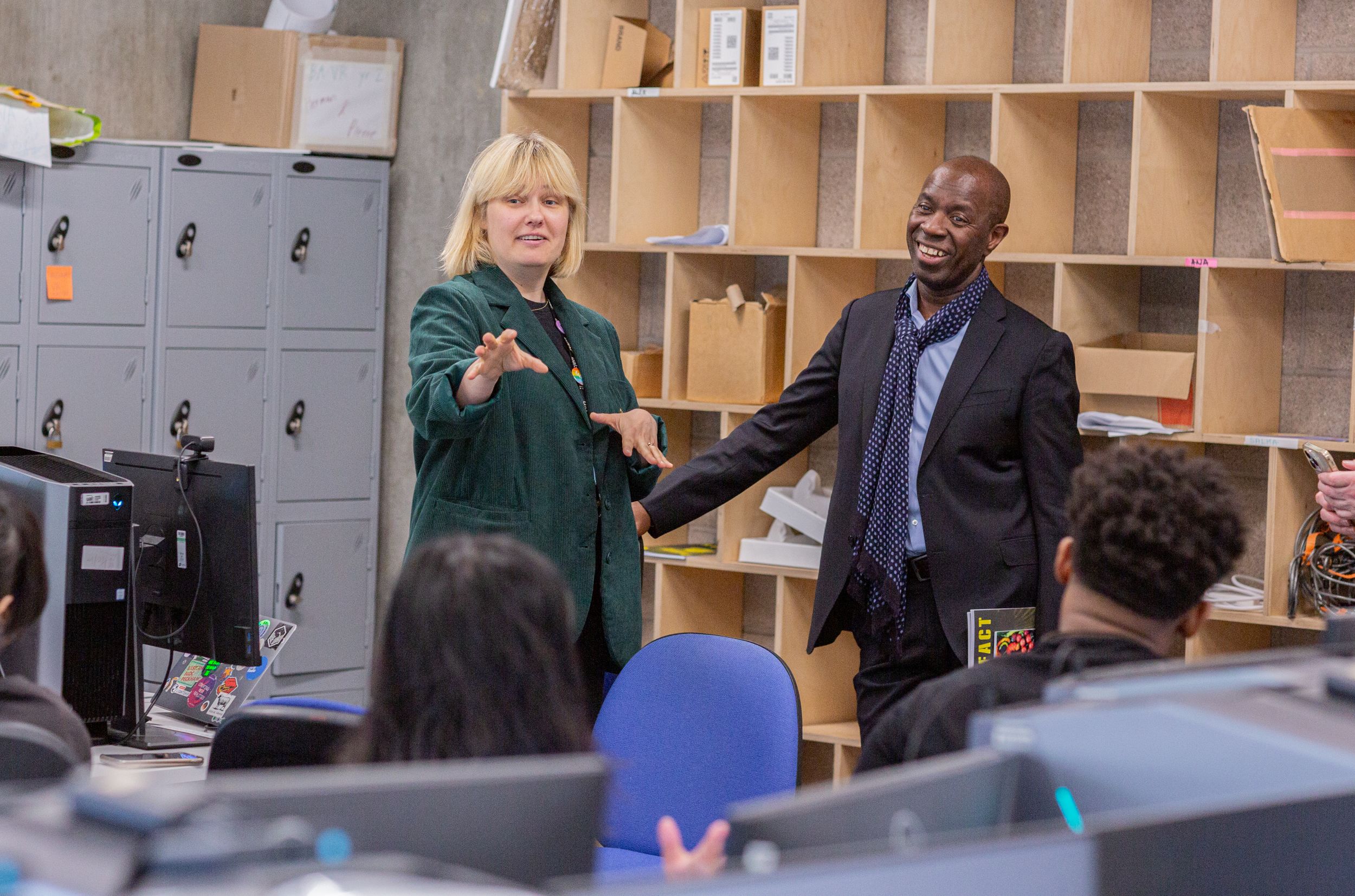 Two people, one woman and one man, are standing and smiling in a workspace with lockers and shelves in the background, while addressing a seated audience. The woman is gesturing with her hand, and the man has an open posture, engaging with the attendees.