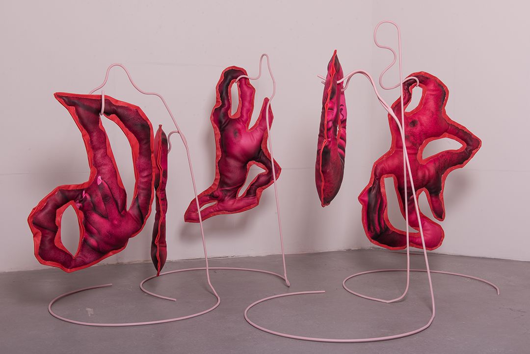 Three red fabric sculptures handing on pink structures. They are situated in a grey room. 
