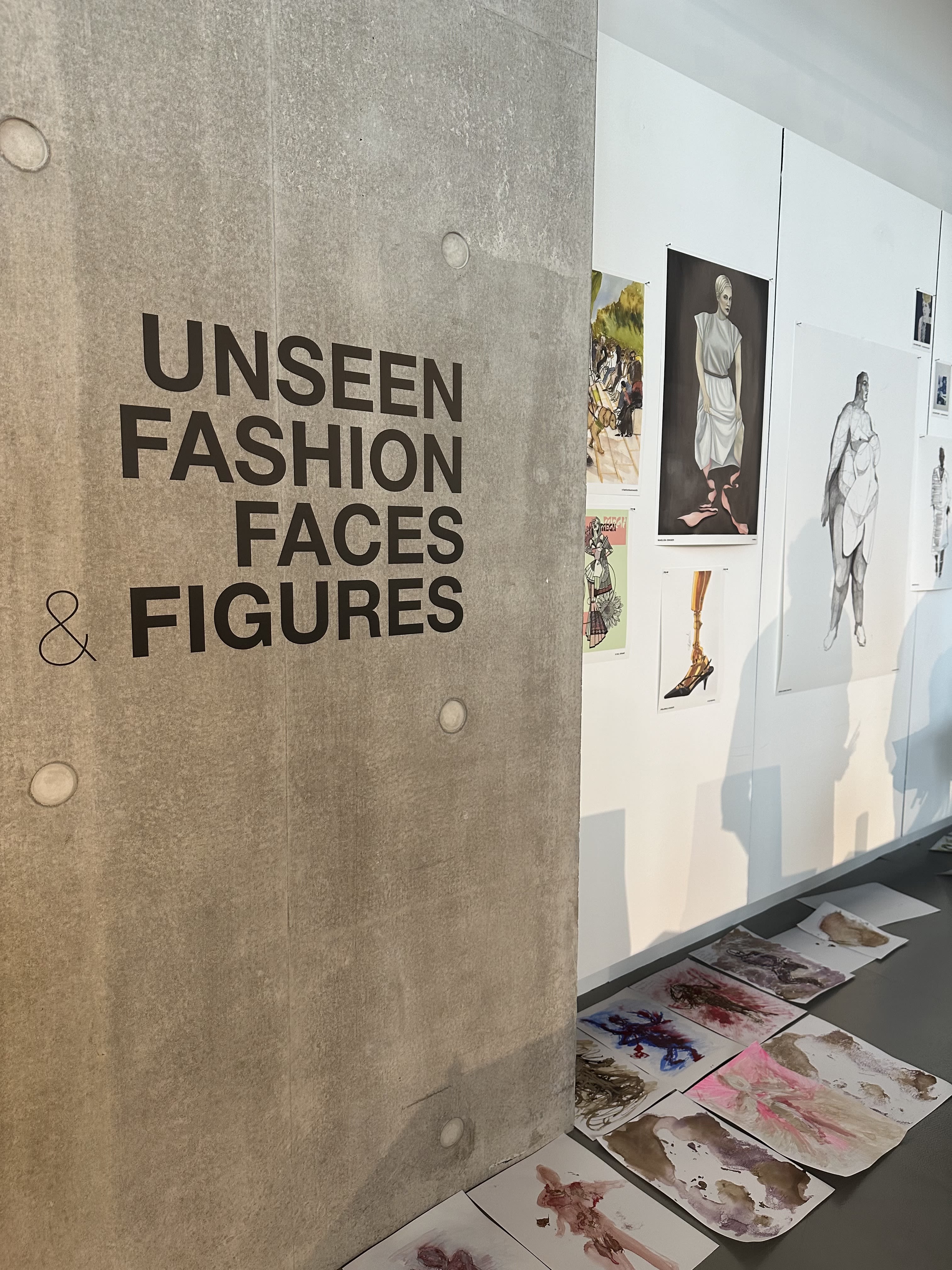 Wall with 'Unseen Fashion Faces & figures' printed on in black, next to white wall with art displayed on it.