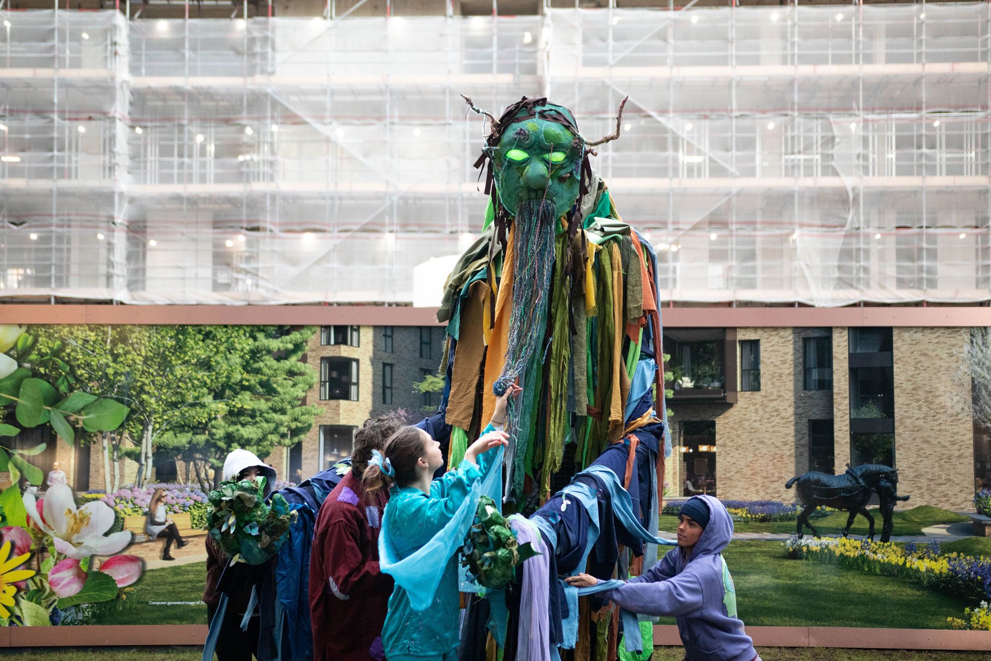A large puppet with a green face, glowing eyes and horns is operated by a group of people wearing costumes in shades of blue, purple and lilac against a backdrop of a hoardig and scaffolding.
