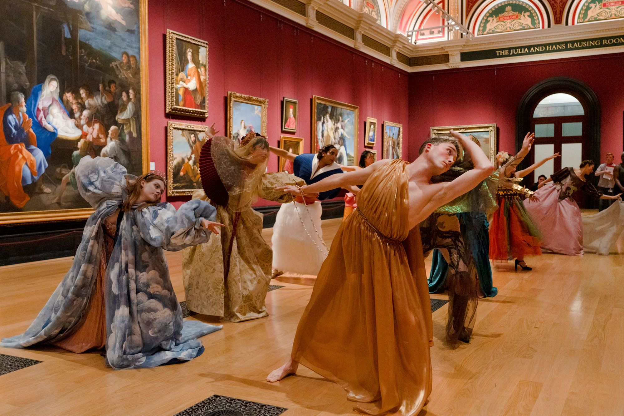 A group of people in different elaborate costumes dance in formation, all with their arm out-stretched, in a large ornate gallery space.