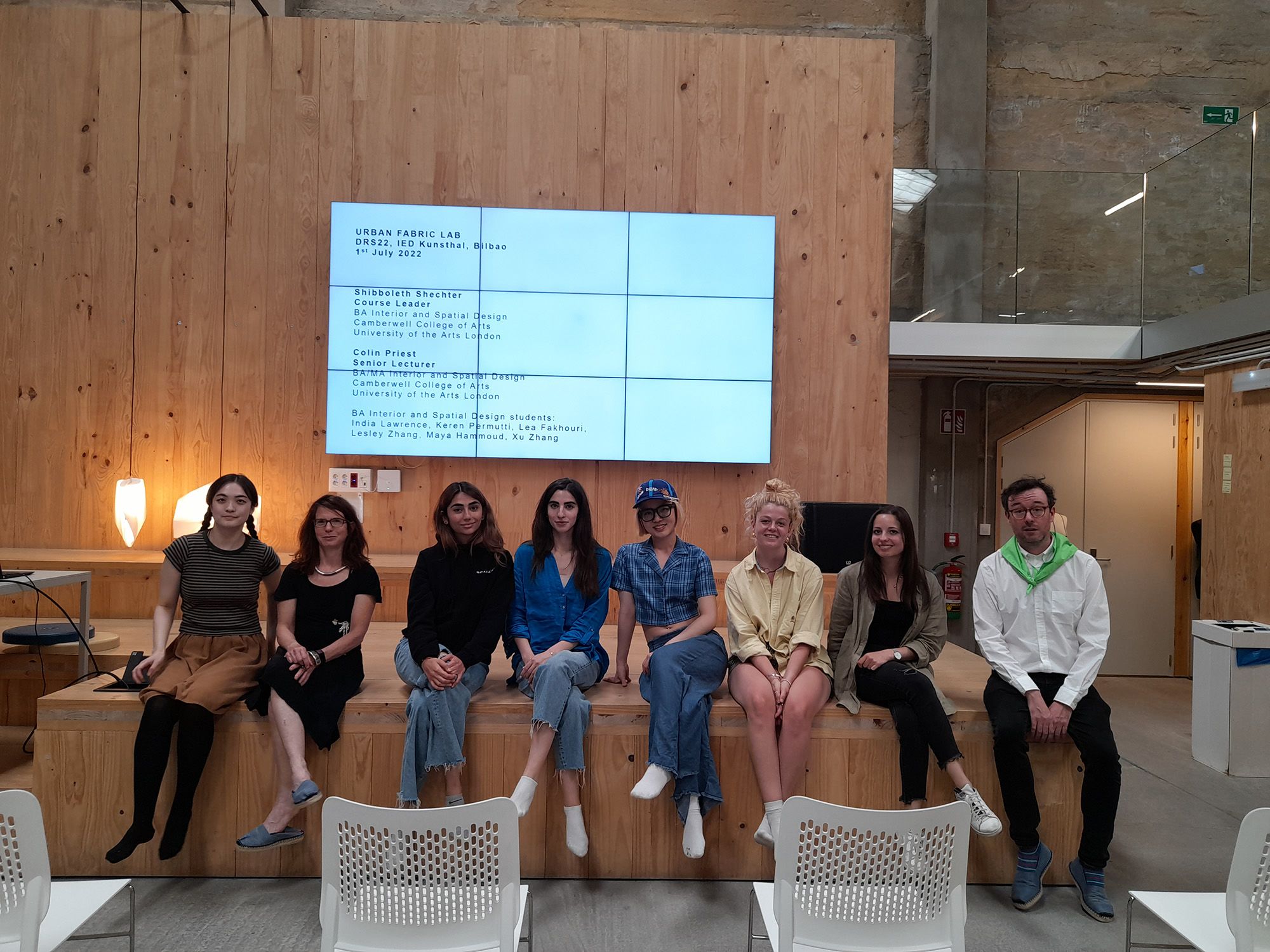 8 people sit on the edge of a wooden stage. They are all facing forward looking towards the camera. Behind them a large screen with the title ‘”Urban Fabric Lab” DRS22, IED Kunsthal, Bilbao, 1st July 2022’. In front of them is a row of empty chairs.