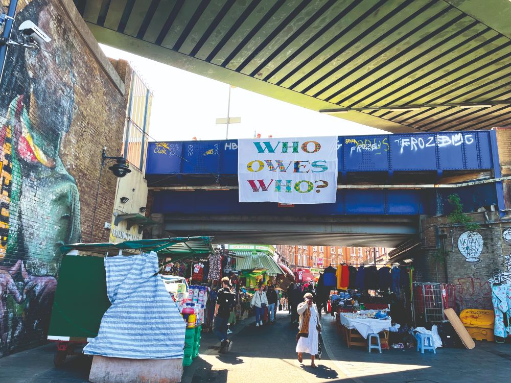A bridge above a market, with a sign on it saying 'Who owes who?'