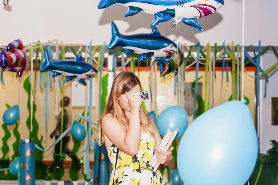 A room full of balloons and a person taking a picture on a point and shoot camera while holding a stack of books.