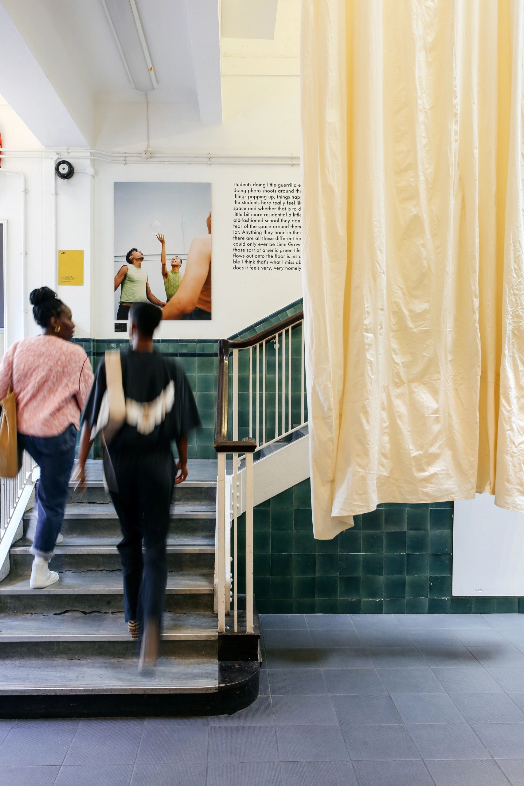 Two students walking up the steps in the reception of UAL Pre-Degree Studies. The walls behind the stairs are made of dark green tiles and a long cream garment hangs from above as an installation piece next to the stairway.