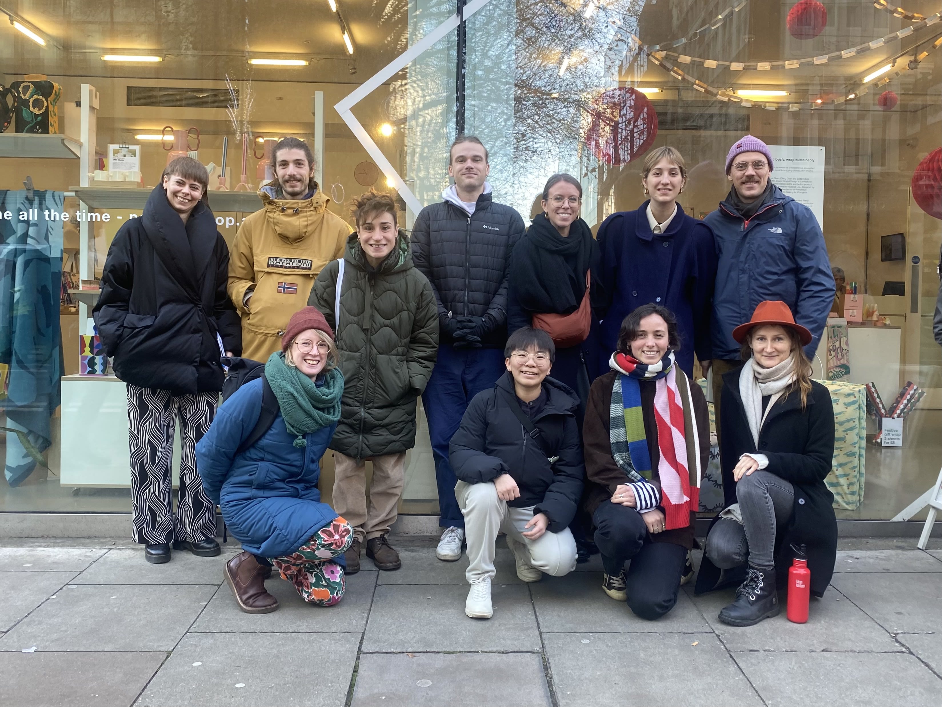 A group of eleven people smiling and posing in front of a shop window with a modern display. The individuals are dressed warmly in winter coats, scarves, and hats, standing on a sidewalk. The shop features creative decorations and text on the window, adding a festive or artistic vibe to the scene.