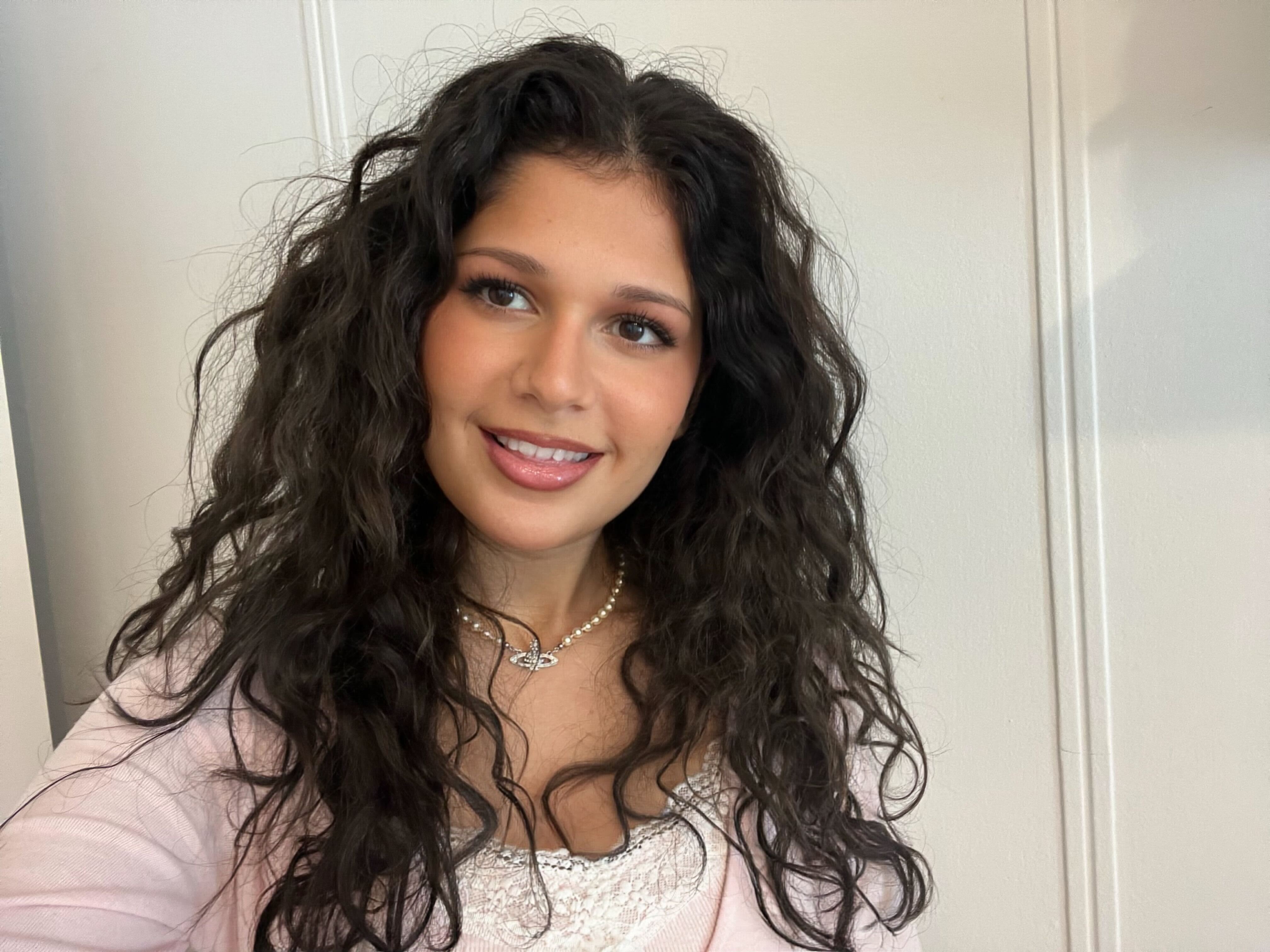 Headshot of Sabrina Plant, smiling and standing in front of a white background, wearing a pink top and necklace.. 