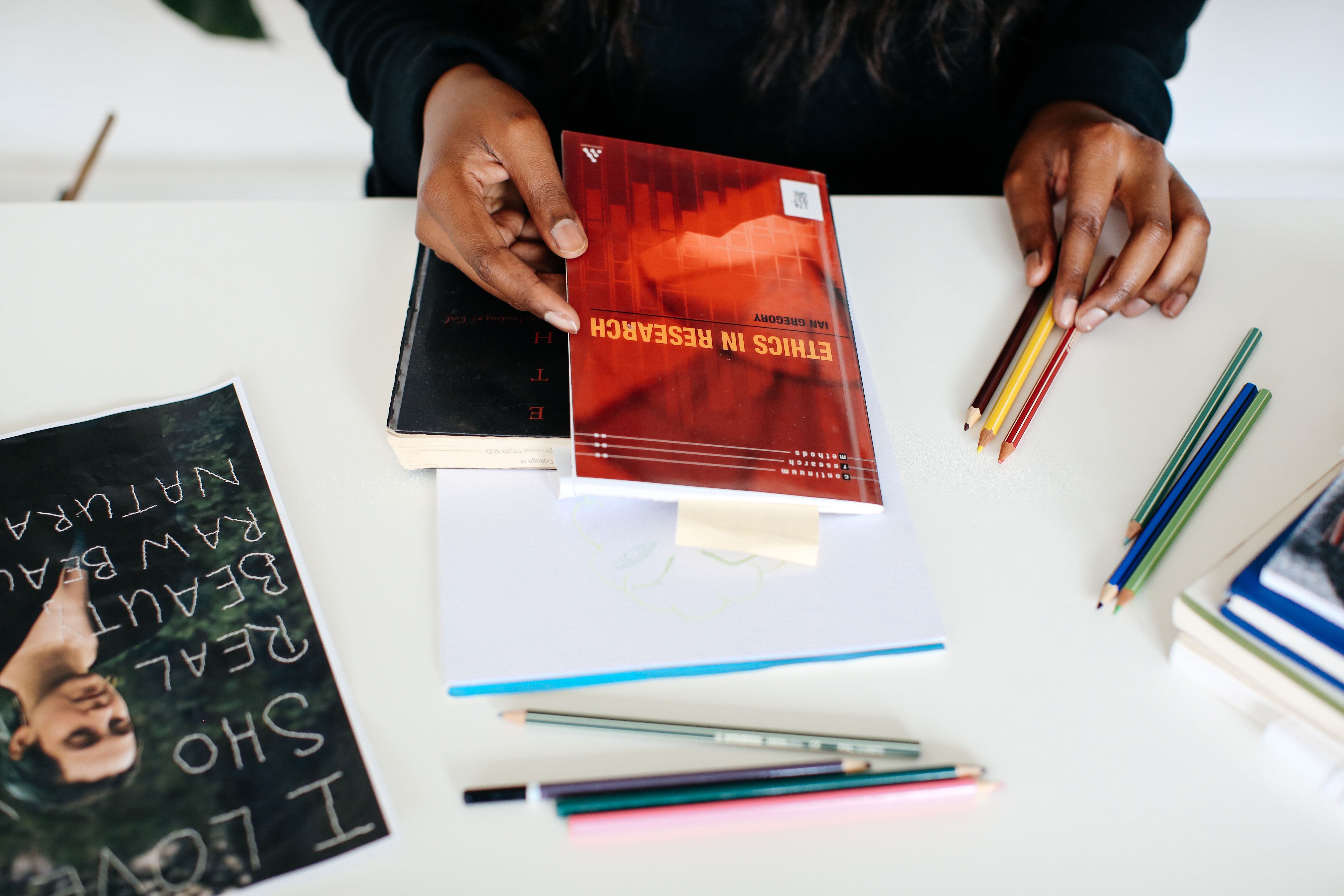 Person's hands holding book which on a table. Colouring pencils on table.