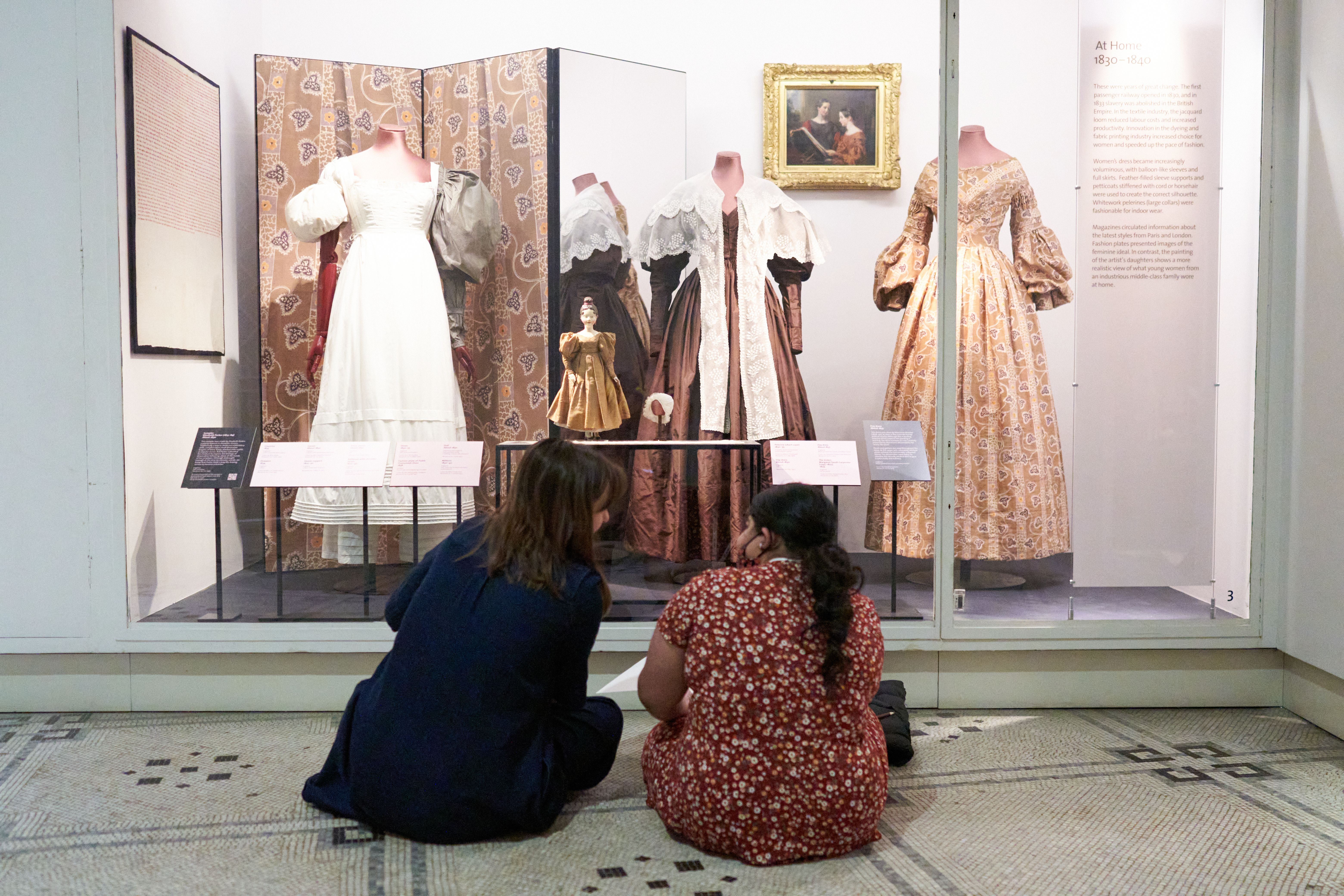 Two people sit on the floor in front of a museum display case containing historic garments, examining fashion objects and exhibition interpretation panels. Photography: © Victoria and Albert Museum, London.