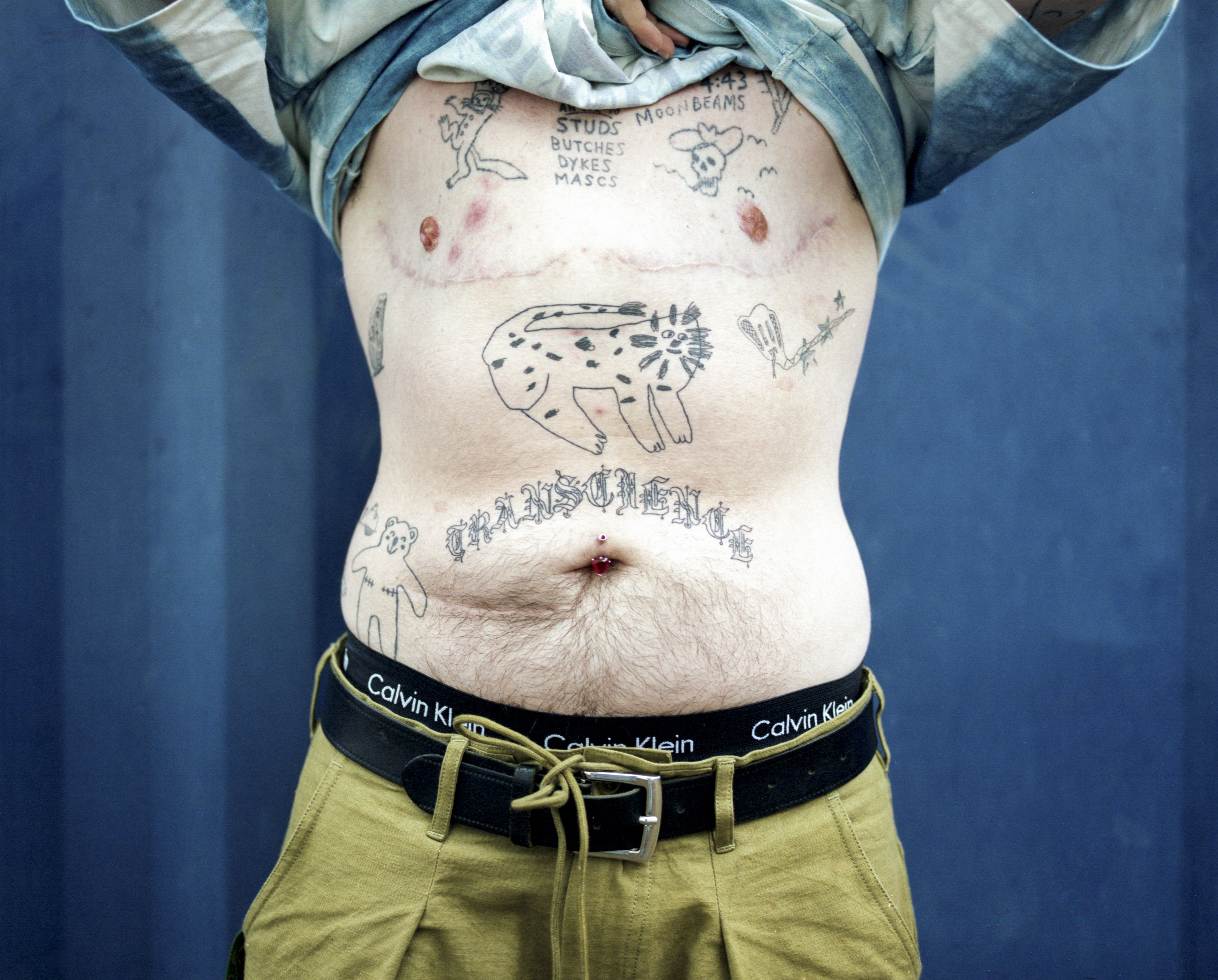 A person raises their shirt to reveal a chest covered in tattoos. The tattoos are of a doodle-like, cartoonish style.