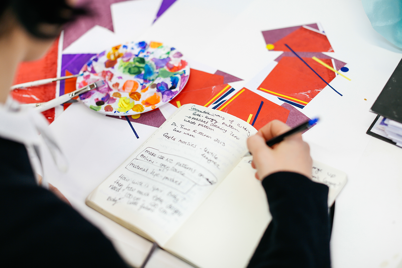 Overhead close-up of someone writing in a journal with colourful papers lying around on the table.