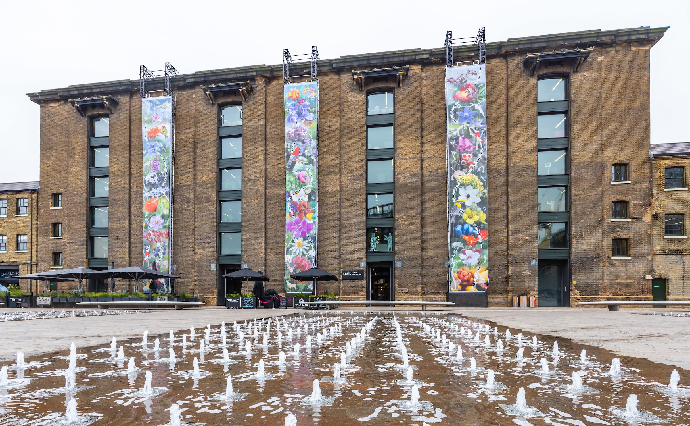 Three ground to roof vertical hanging botanical prints hanging on the front of the Central Saint Martins Granary Building, with the fountains in front visible