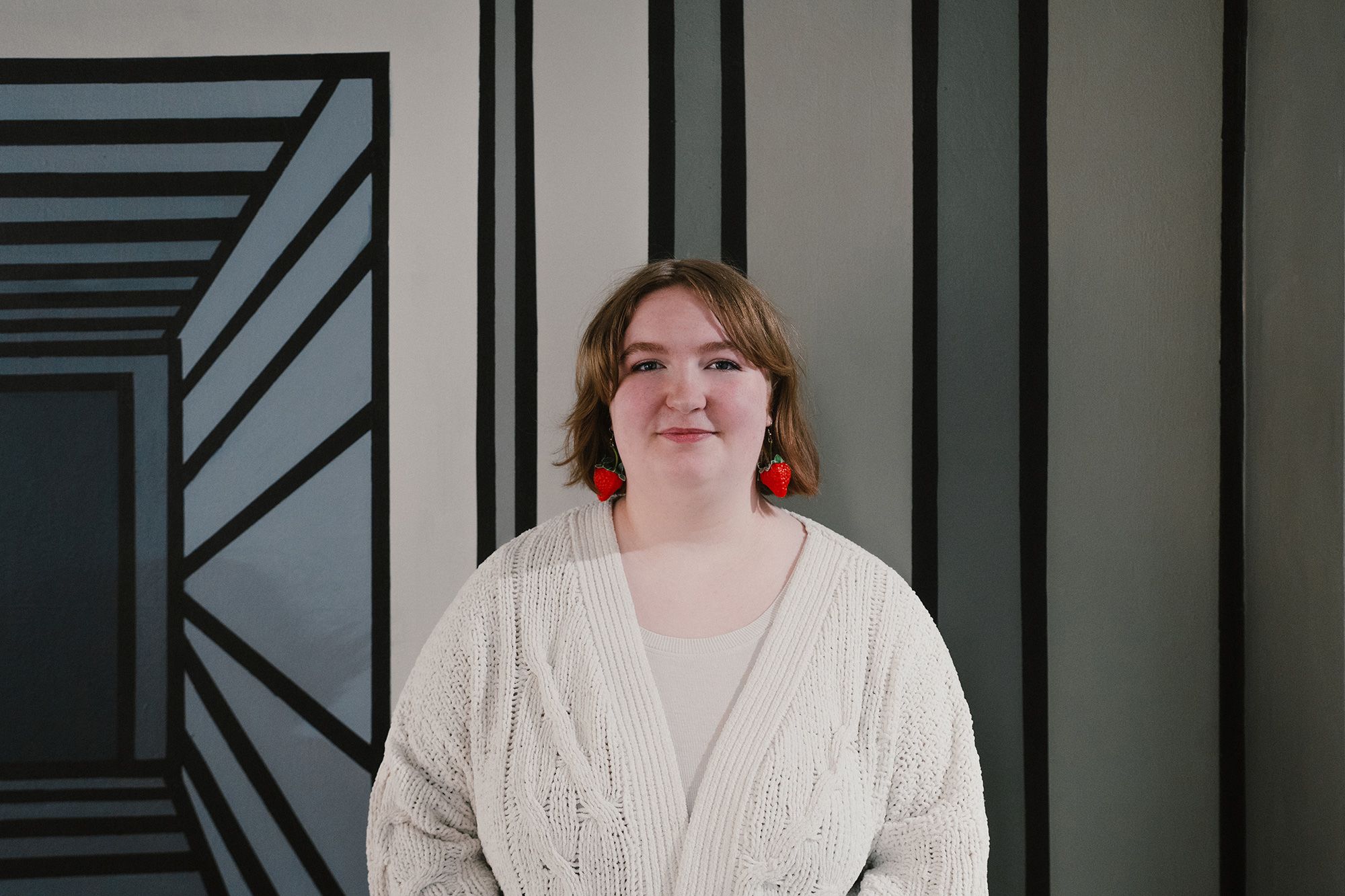 Image shows a person wearing a white cardigan and small red earrings with short brown hair smiling while looking at the camera. In the background there is a painting that uses different shades of grey.