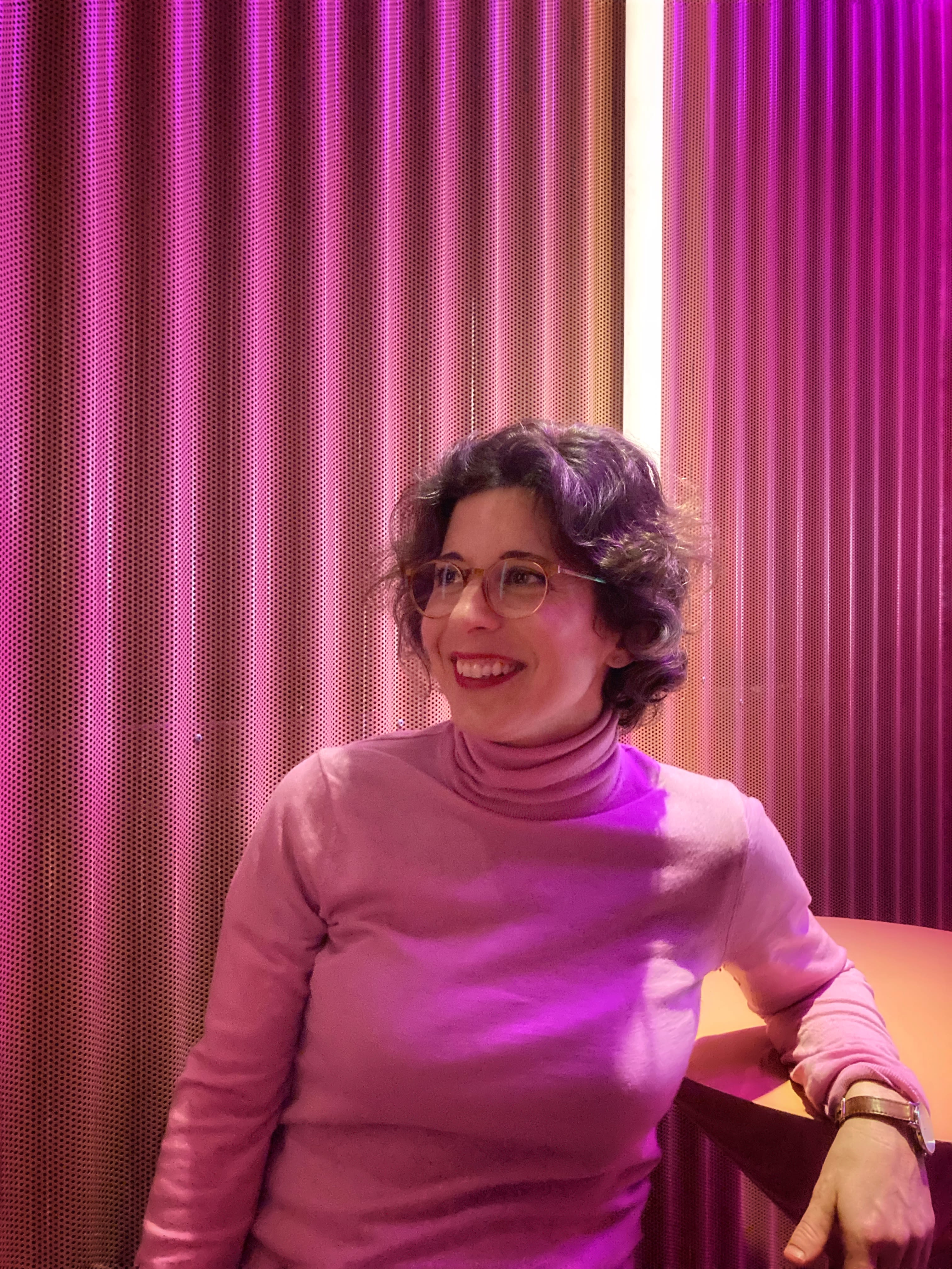A white woman with short curly hair and glasses seated in front of some drapes. She is smiling and looking to her left.