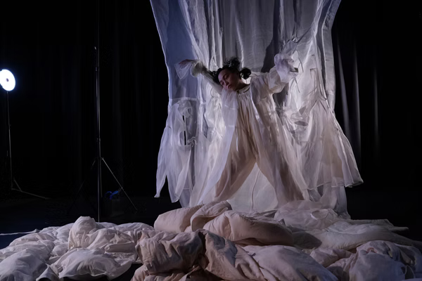 A performer suspended in flowing, sheer fabric above a soft, padded floor in a dark studio, lit dramatically from the side, creating a dreamlike, floating effect.