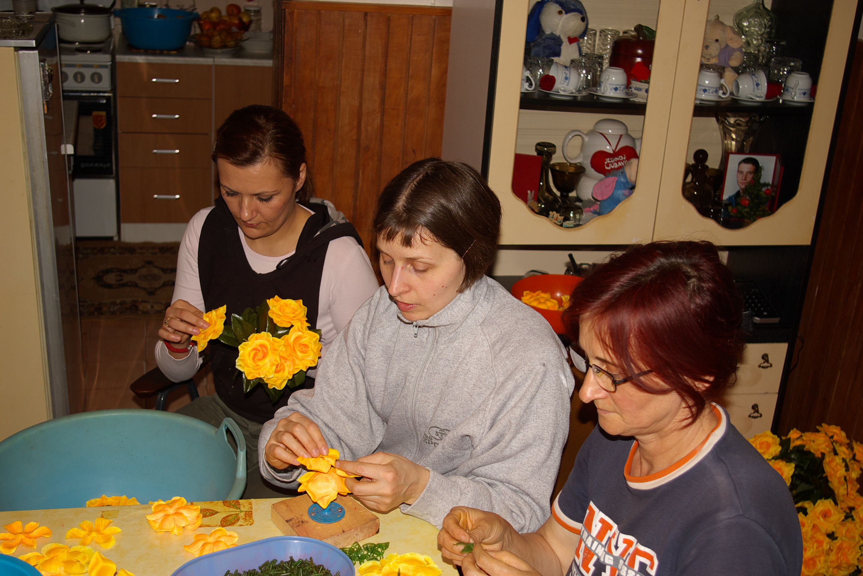 3 individuals crafting with flowers at a table in a domestic setting, with a sideboard and room in the background