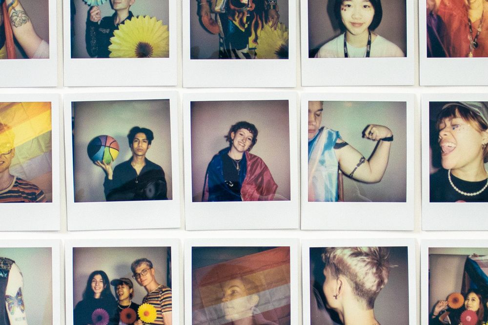 A wall of polaroids, showing different members of the UAL LGBTQ+ community. Emma Martin is in the centre, with a Bi flag draped around her shoulders.
