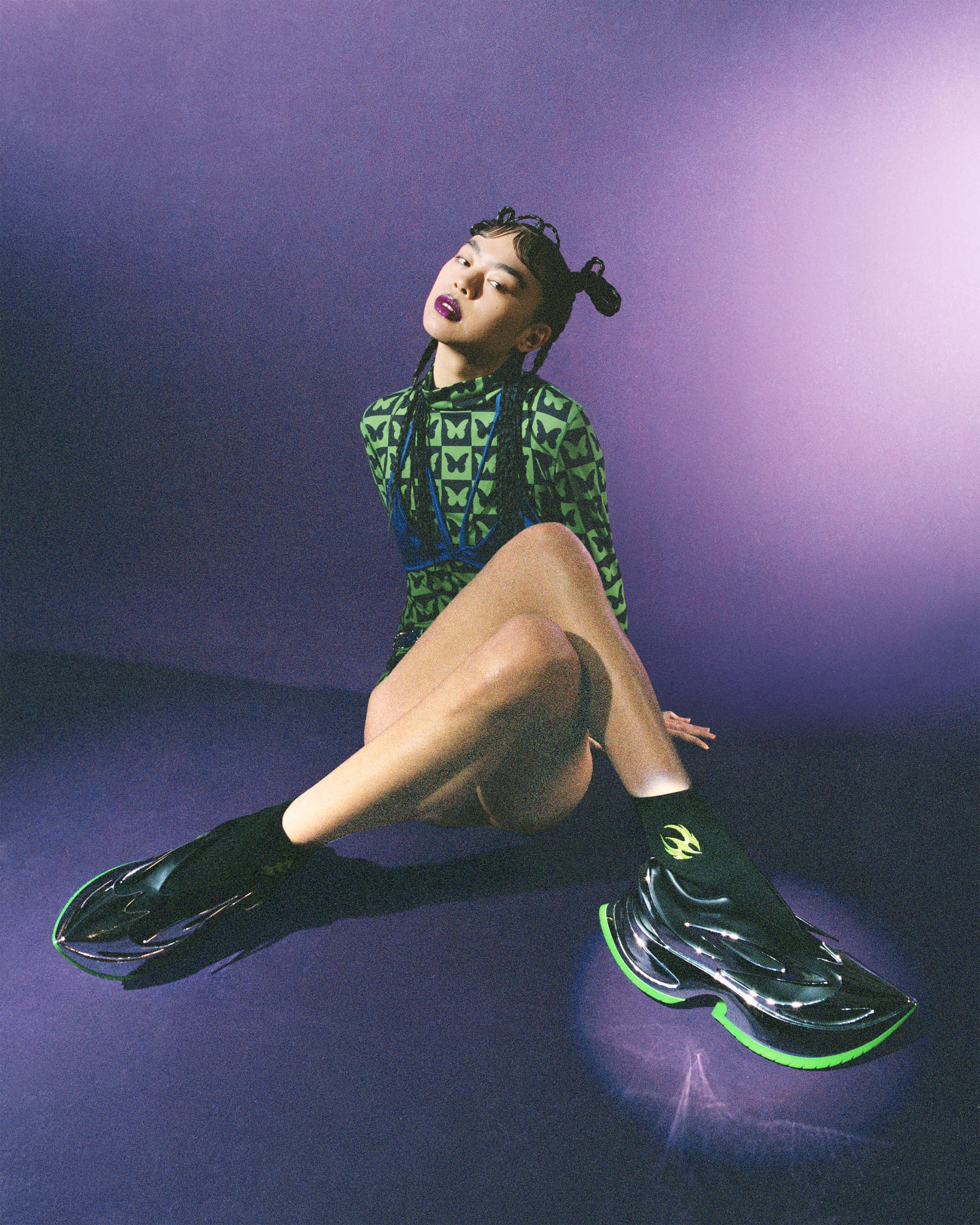A person sitting on the floor, the background is purple and they are stretching out their leg to show a black and neon green trainer