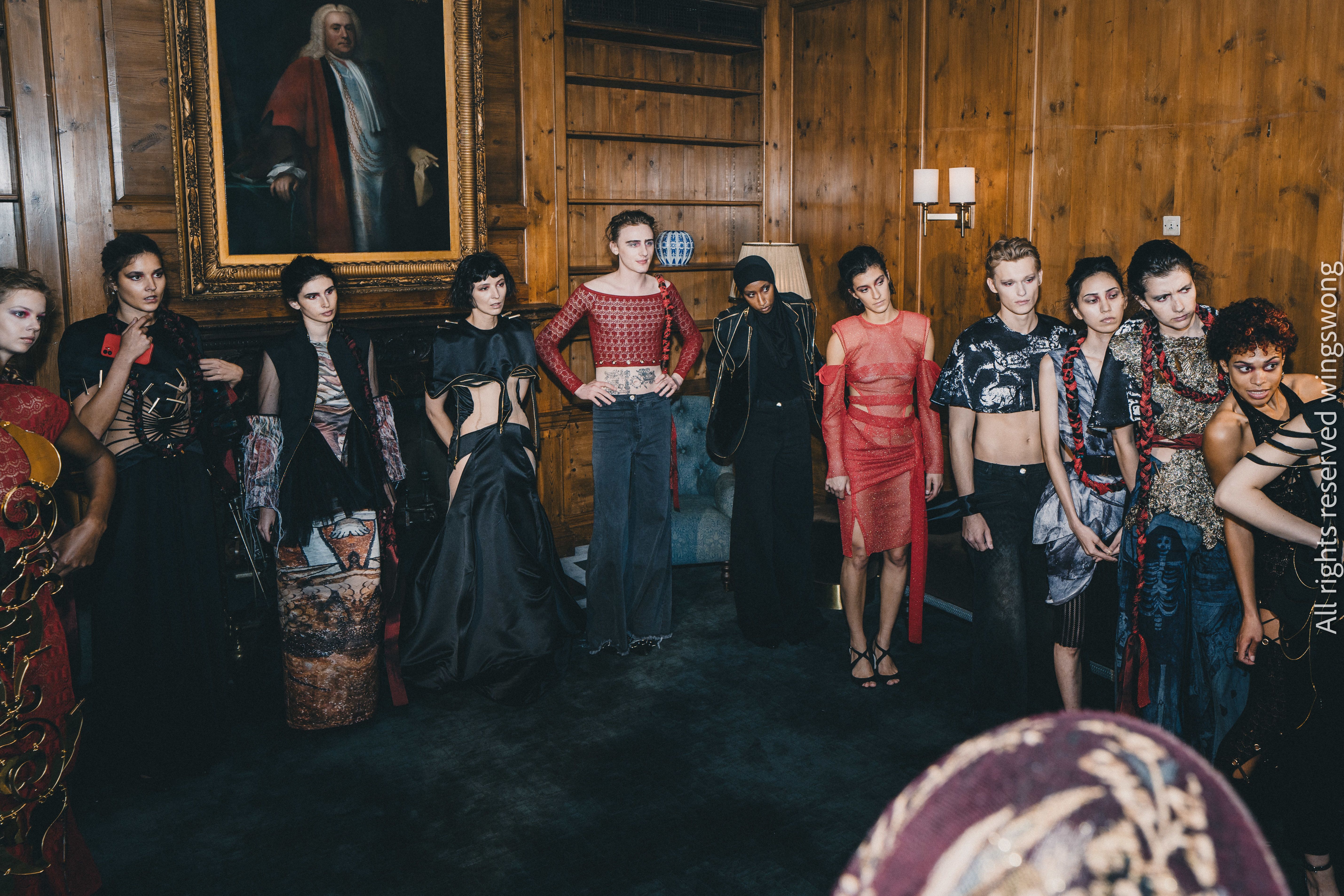 A diverse group of fashion models in eclectic outfits stands in a dimly lit room with wooden walls and a classic portrait. The mood is edgy and artistic.