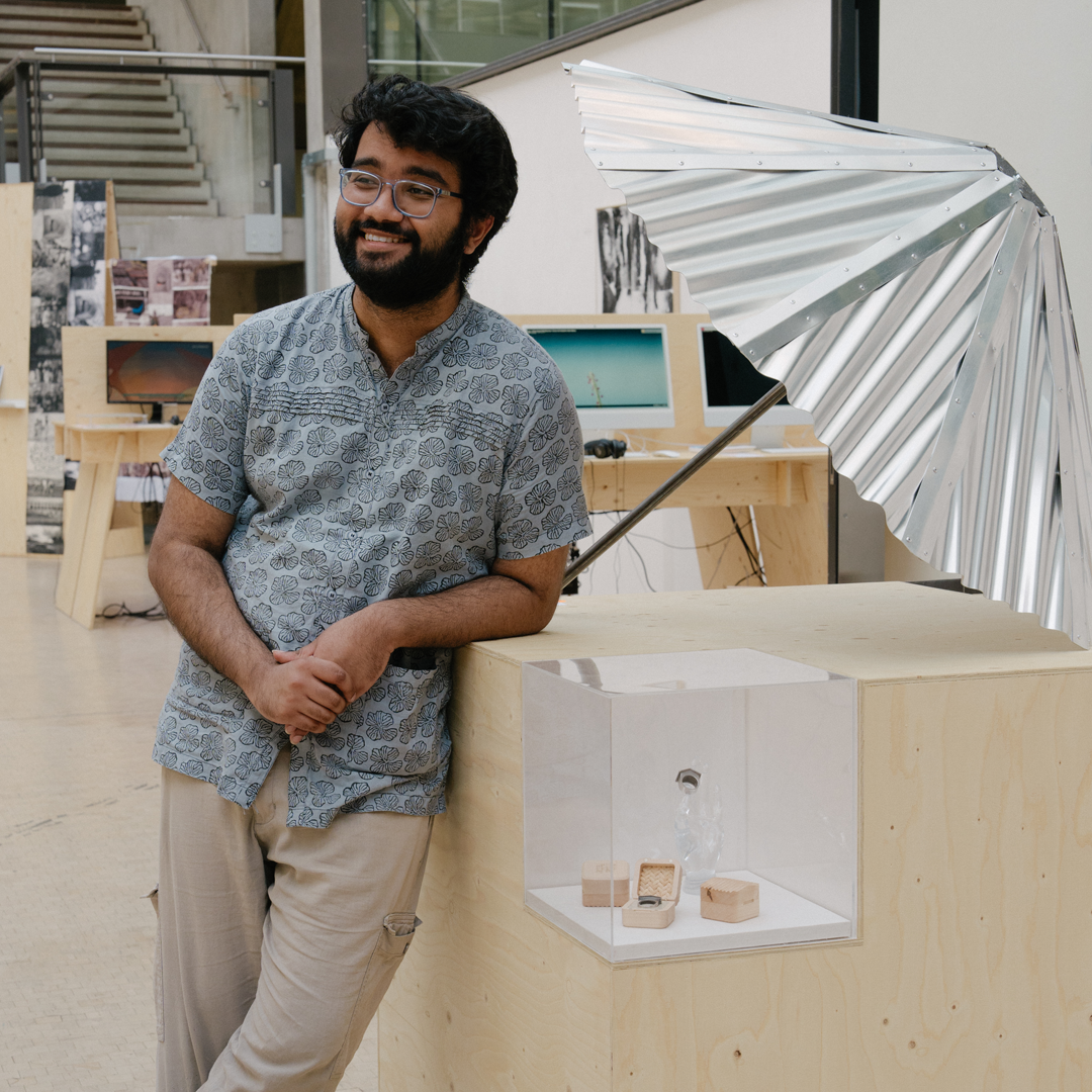 A student stands smiling in a studio, leaning against a wooden display plinth with a metal umbrella installation on top, surrounded by desks and creative materials.