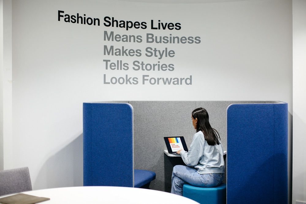A student on their laptop in a working cubicle, sitting below a sign reading 'Fashion Shapes Lives'