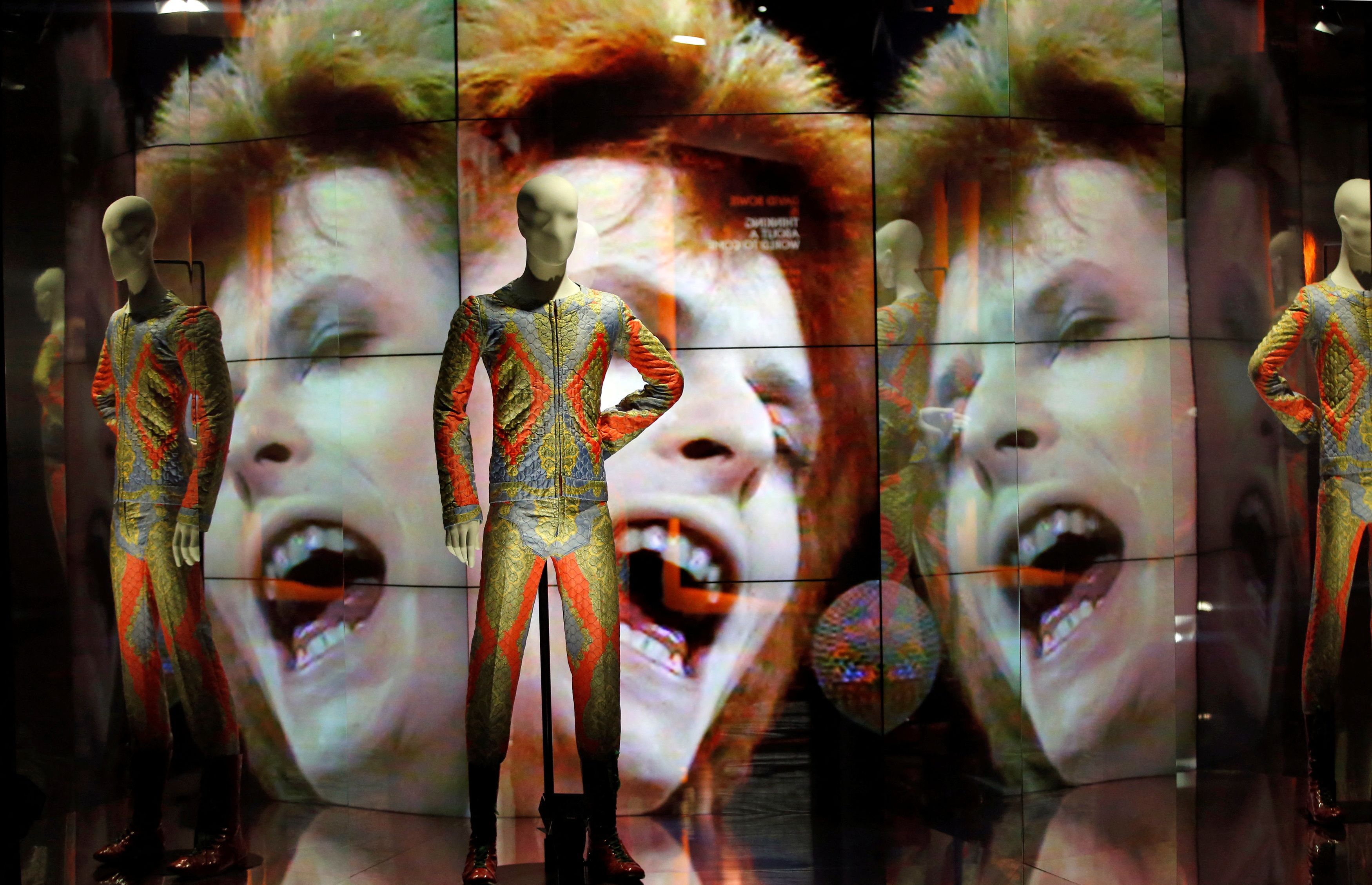 Two suits designed by Freddie Burretti (1972) for the Ziggy Stardust tour are displayed in front of a video showing pop star David Bowie singing at the BBC show Top of the Pops (July 1972) as part of the exhibition 