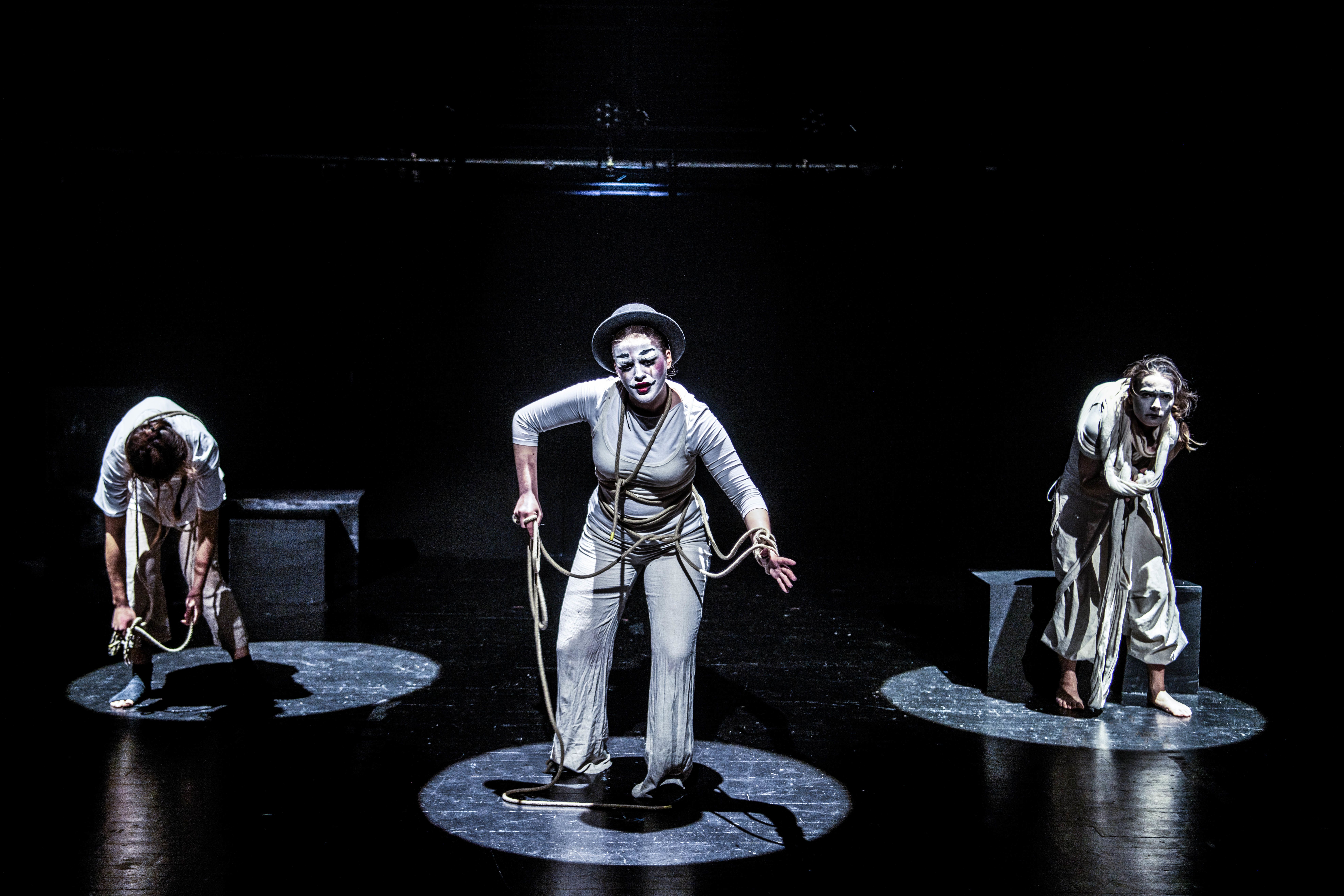 Image shows 3 people in white spotlights standing on a darkened stage. The person in the middle has white mime makeup with ropes round them on while the people at either side are bent over also with ropes around them and wearing white. 