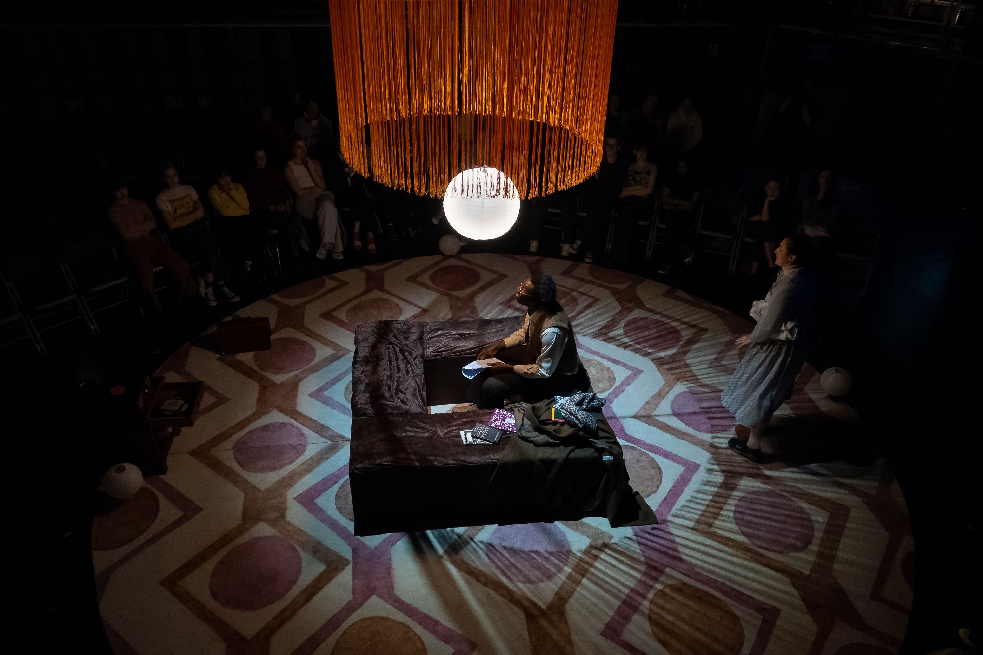 A man is kneeling on a square plinth in the middle of the stage. He is lit from above, and in the shadows around him stands a circle of people. The light orb and orange fringing hang from above.