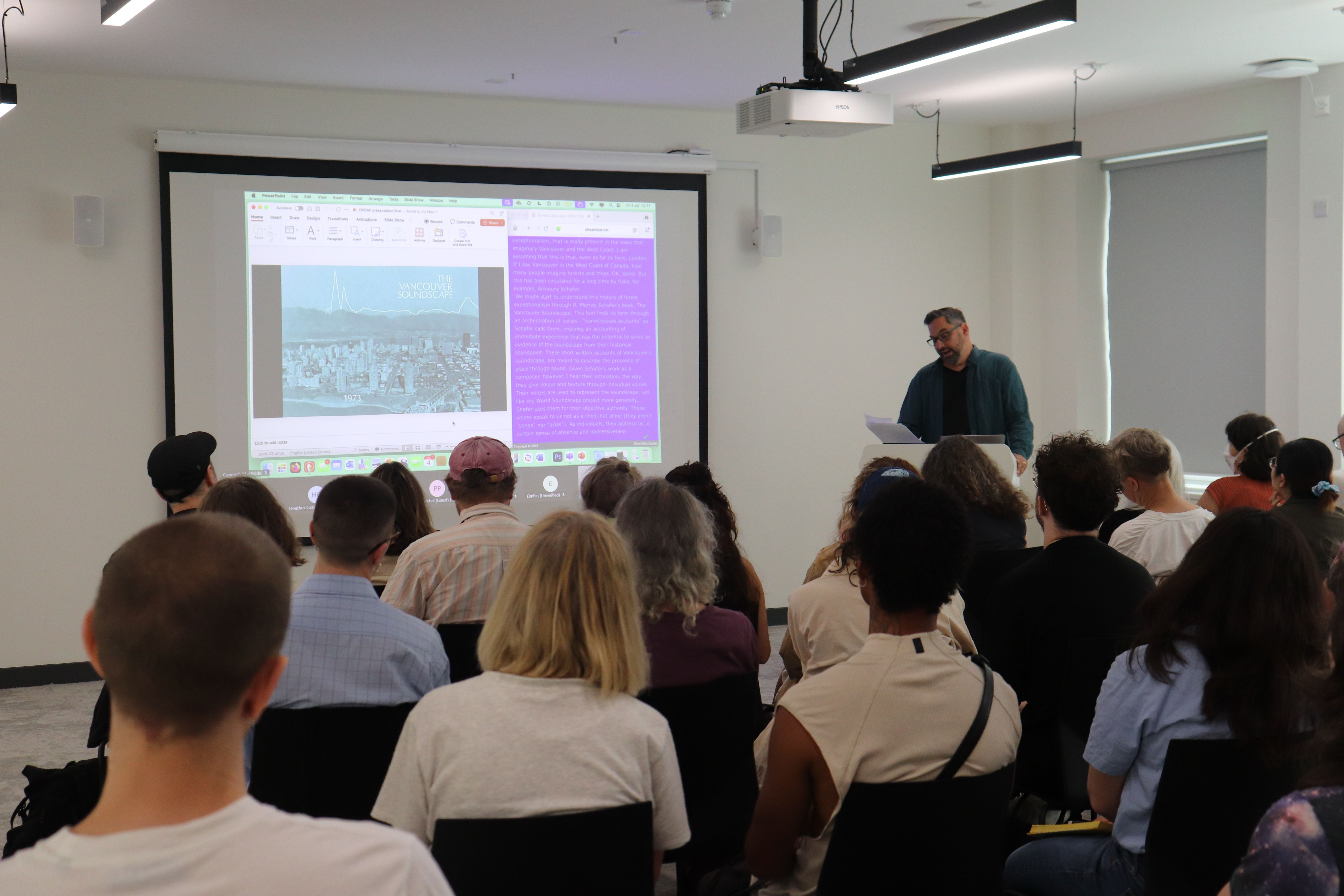 Dylan Robinson by projected slides at lectern reading from paper. Backs of audience heads.