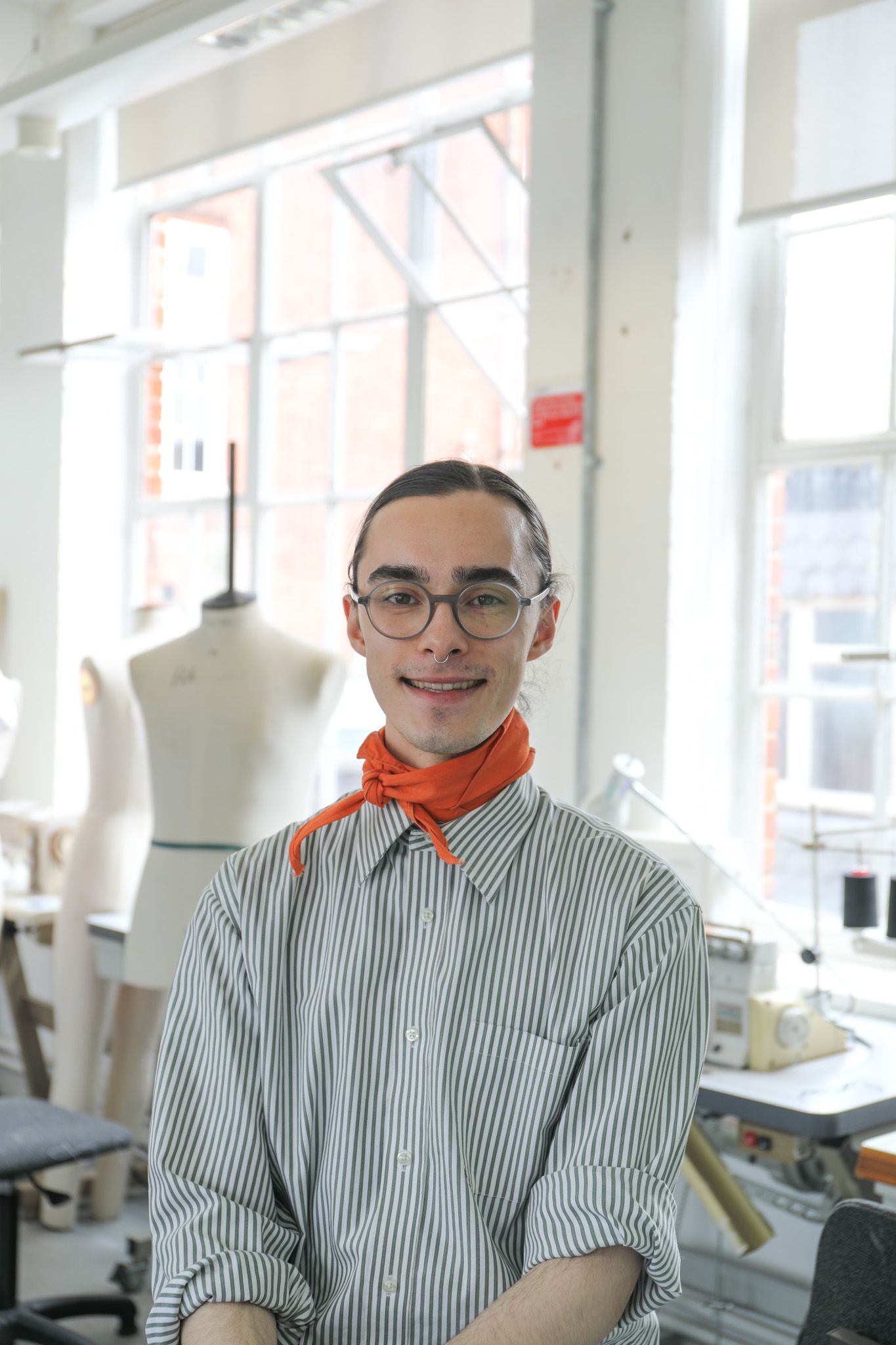 A person wearing a striped shirt and an orange neck scarf sits in a bright fashion studio, surrounded by dress forms, sewing equipment and large windows.