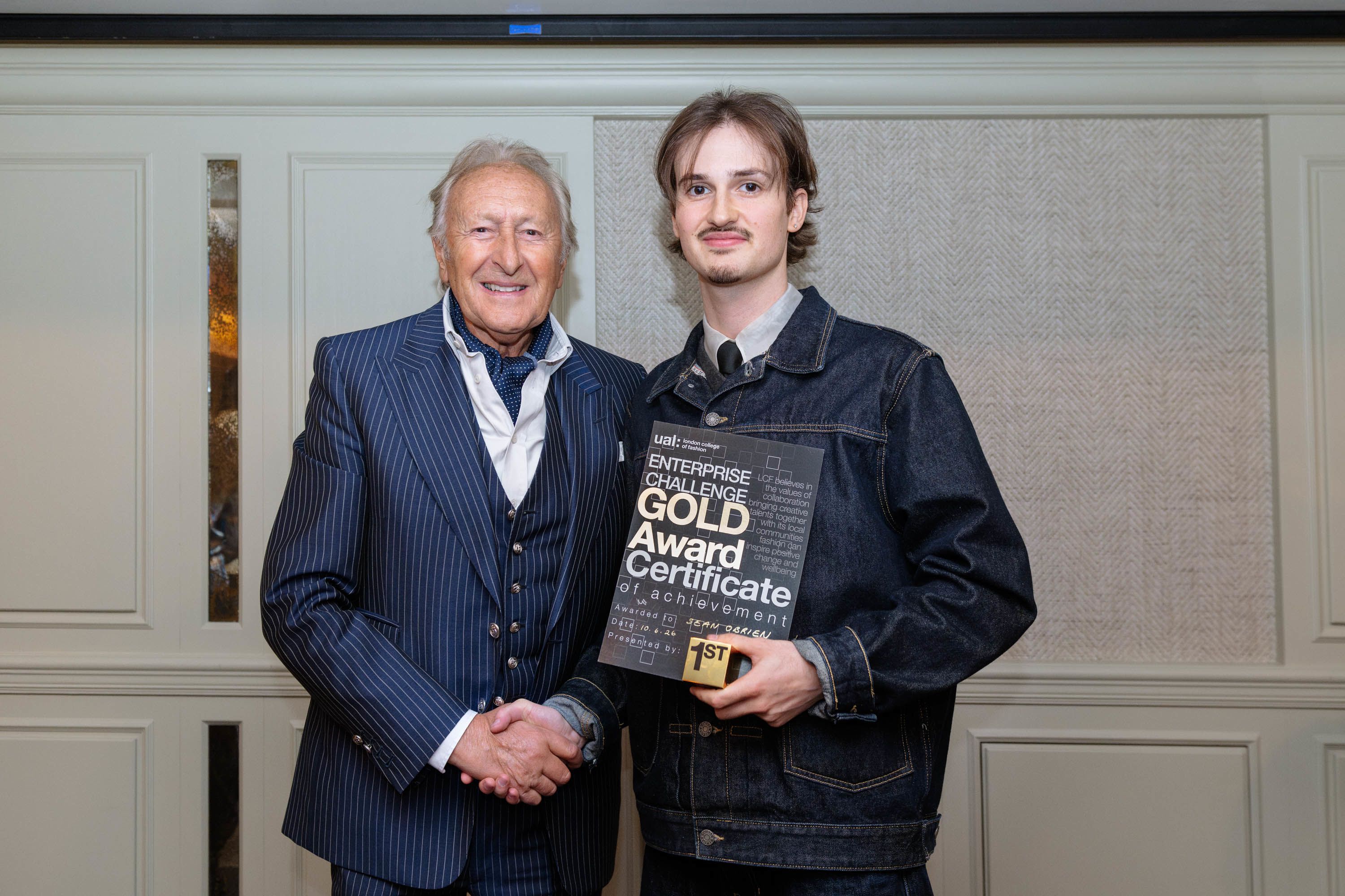 Two men shaking hands on with grey hair and a navy blue pinstriped suit and the other brown hair and navy blue jeans jacket holding a certificate and award.