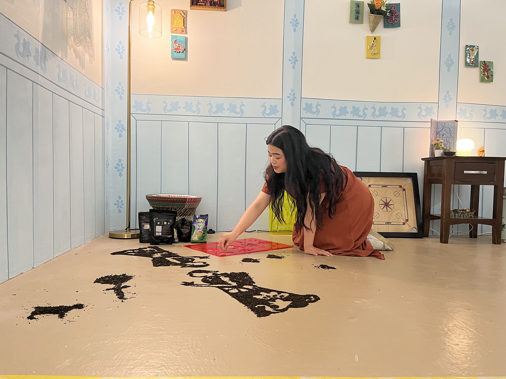 Synchar is on her hands and knees within her installation, on the floor is a red stencil, and shapes made with tea powder. Synchar holds her hand above the stencil and sprinkling tea powder.  