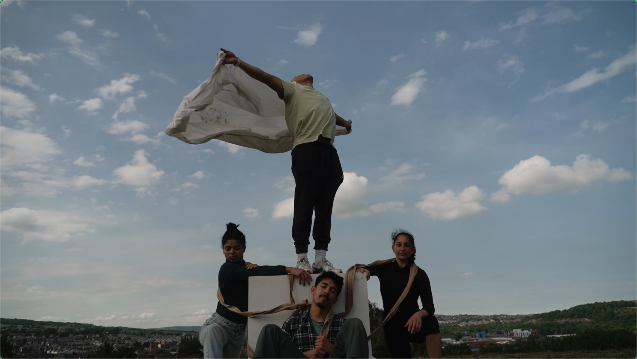 An outdoor film scene of 4 people shot against a blue sky on a hillside. There is a town in the background. They are performing in pose.