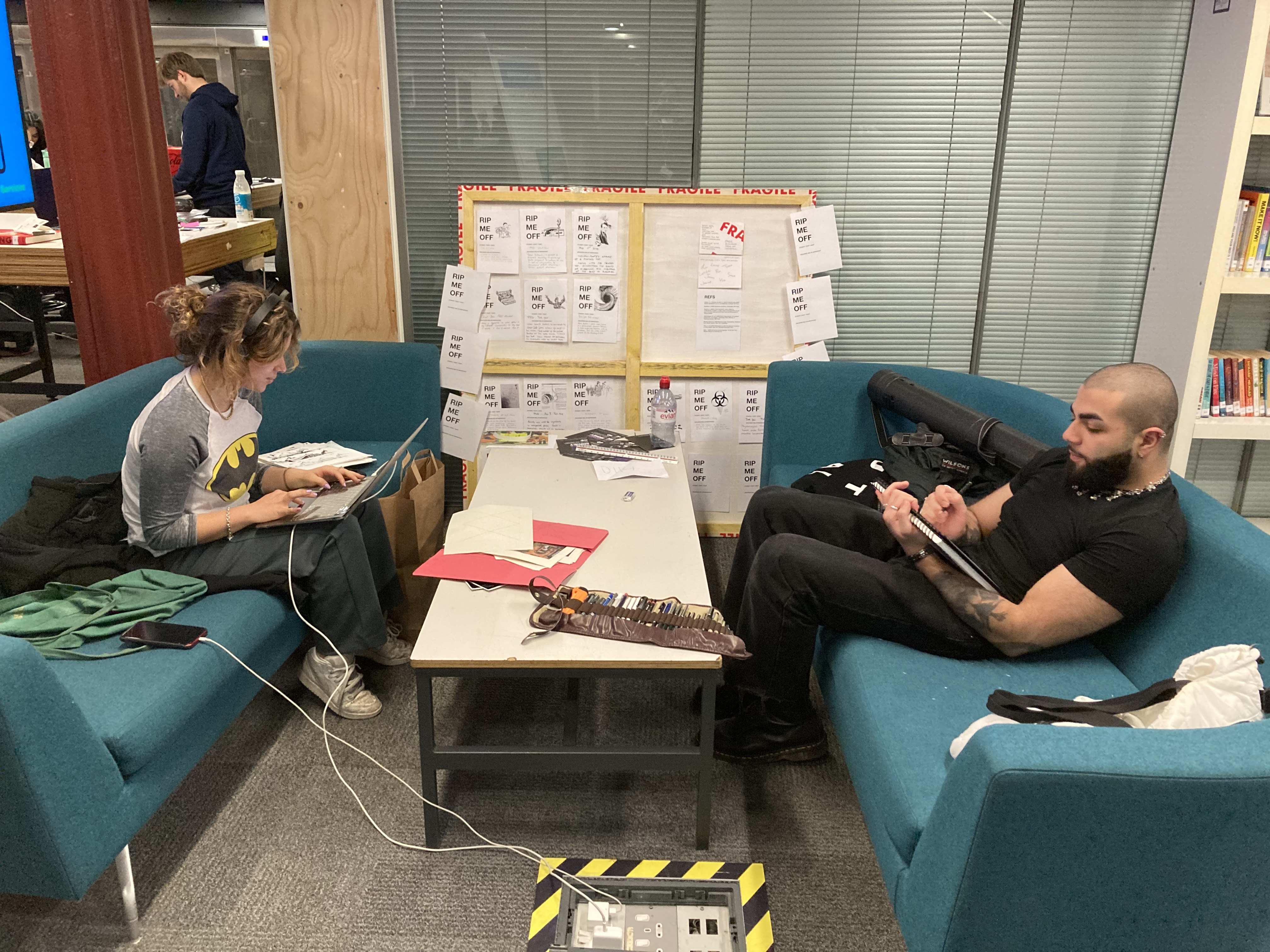 Two people sitting on opposite sofas working on laptops, with a table in the middle.