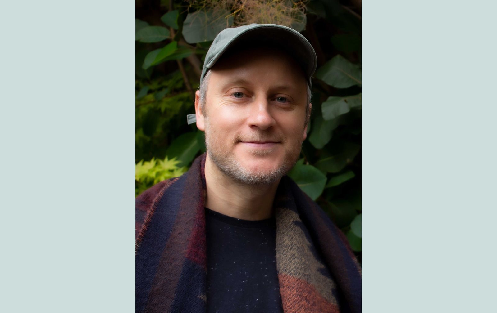 A portrait photo of Mark, smiling to the camera stood in front of a plants, wearing a cap and scarf