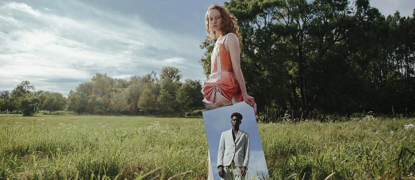A women standing in a field surrounded by trees and grass holding a mirror reflecting the image of a man wearing a suit. Photography by Cottonbro Studio, Pexel.