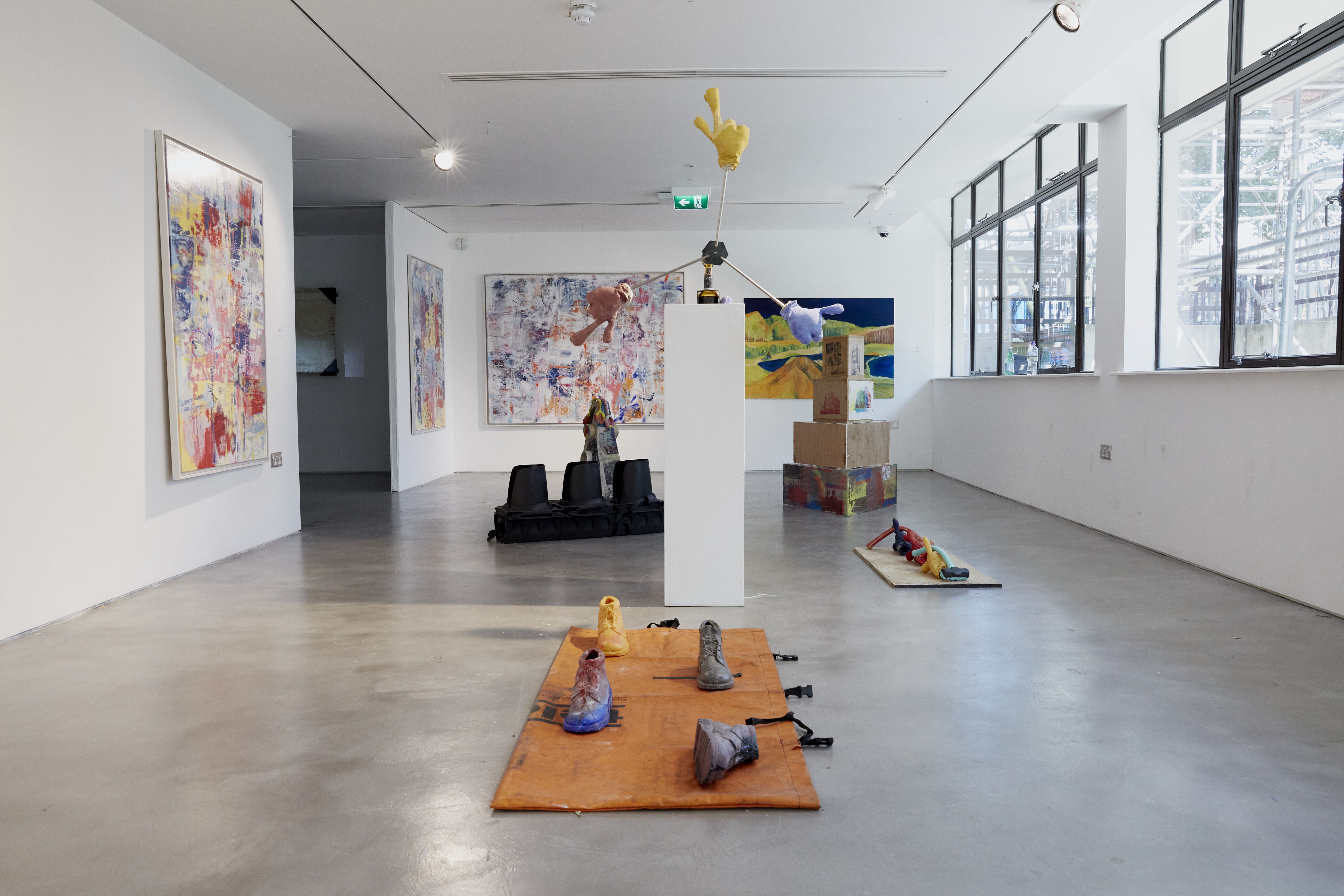 This image shows a contemporary art gallery space featuring various sculptures and abstract artworks. The sculptures include vibrant, unconventional forms displayed on the floor and pedestals, with painted canvases on the walls. The large windows to the right let in natural light, enhancing the clean, minimalist gallery atmosphere