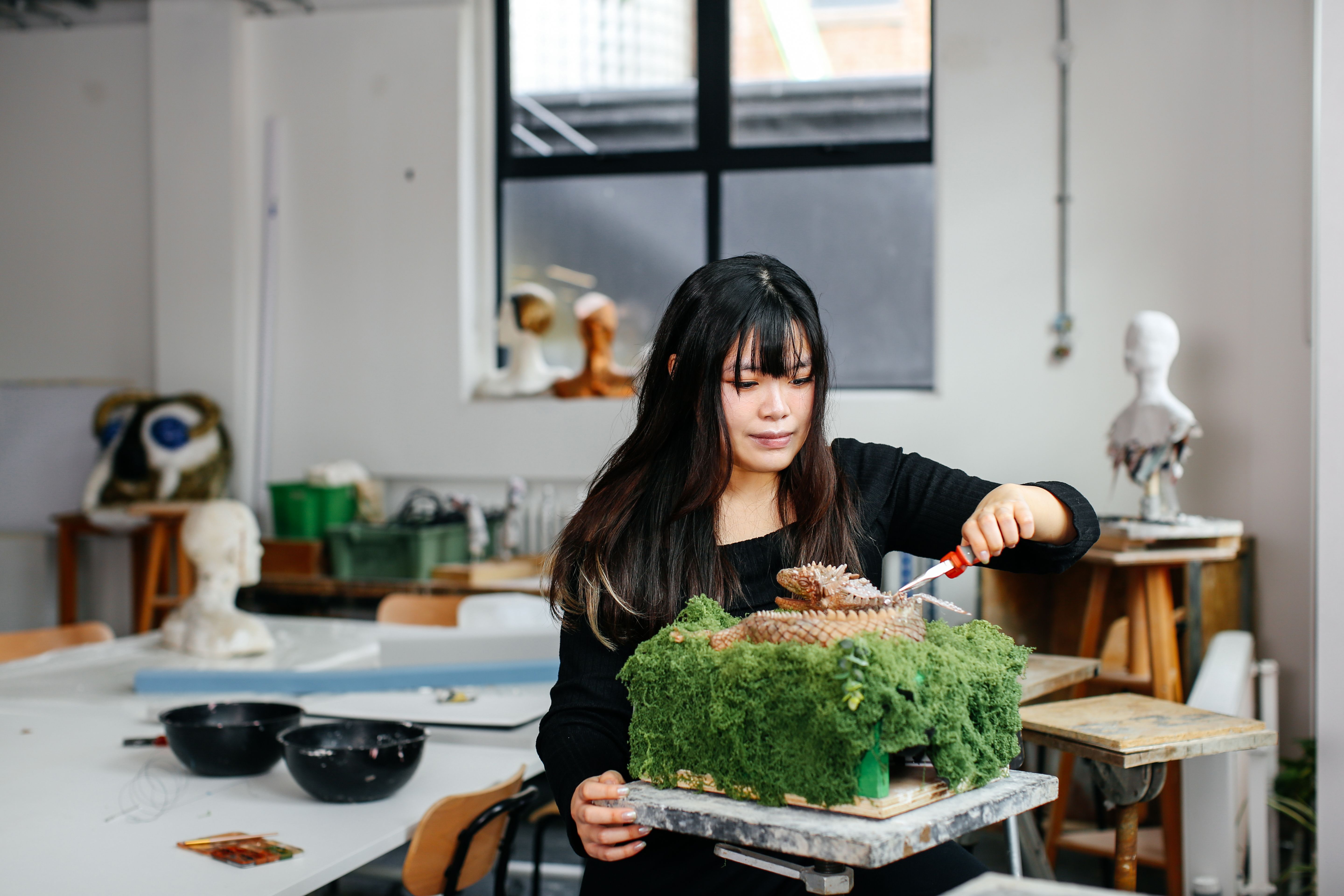 Faye Lok works on a model of her installation Ladon’s Feast in a studio, sculpting a small dragon emerging from a moss-covered forest base.